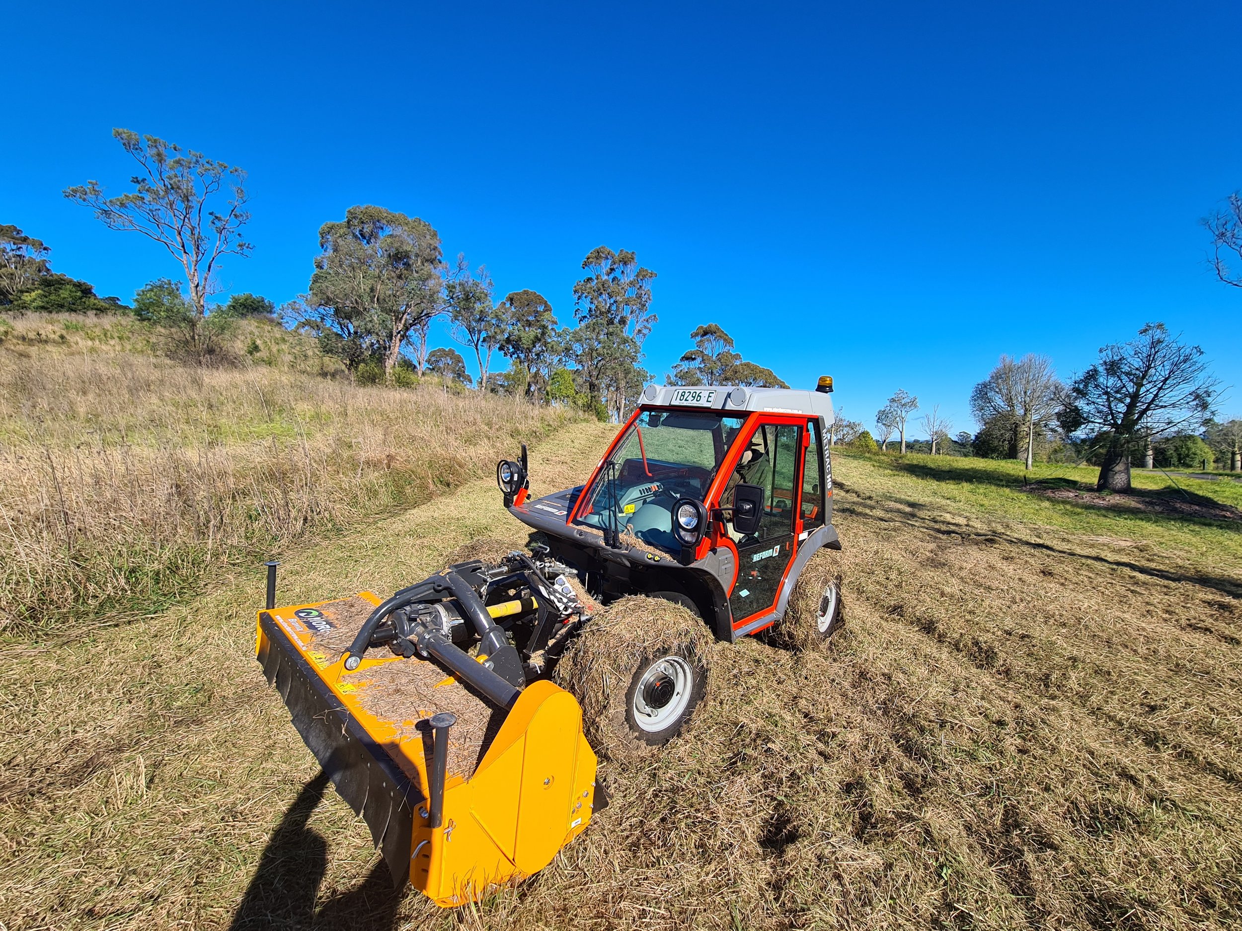 Mower tractor in a grassy field with trees and a clear blue sky.