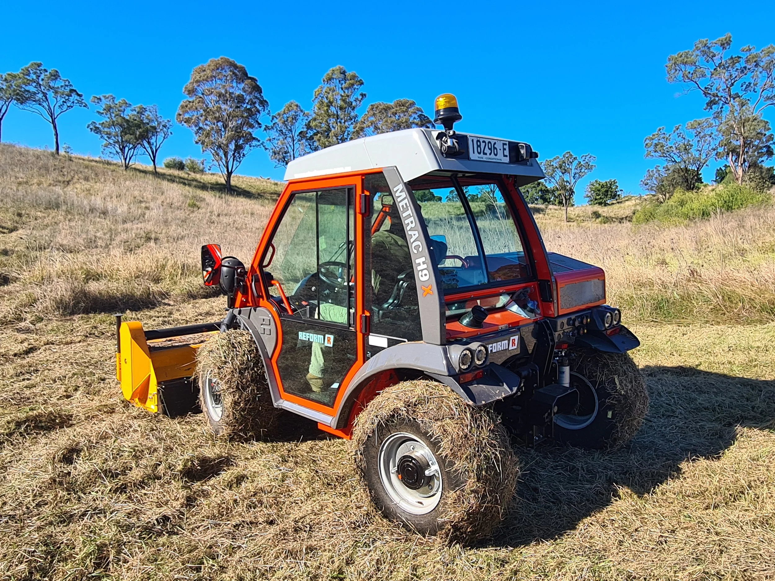 Red and gray tractor with a glass cabin parked on a grassy field under a clear blue sky, surrounded by scattered trees.