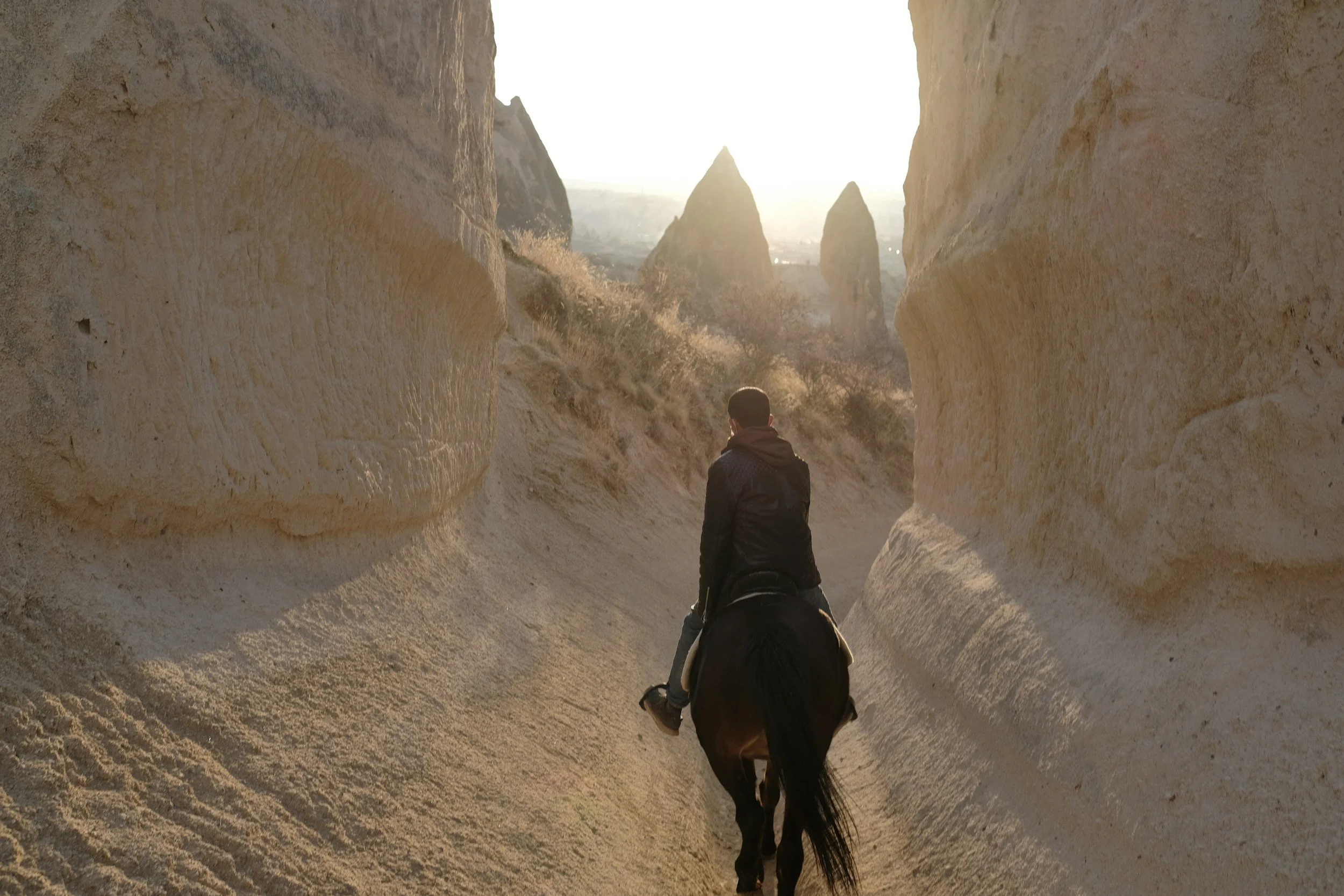 A person riding a horse through a narrow canyon with rocky walls, with tall rock formations visible in the distance and sunlight shining from behind.