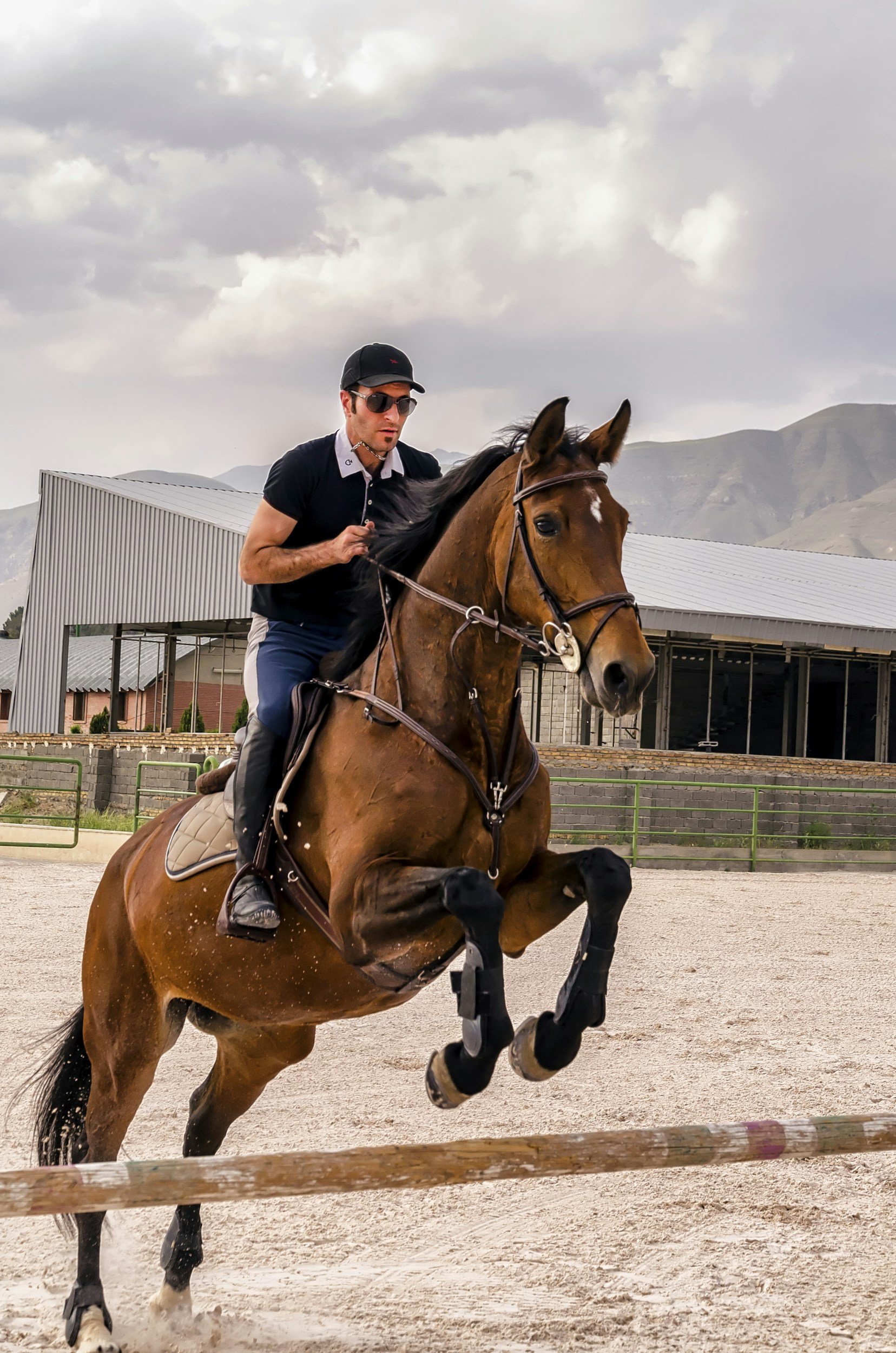 A man riding a brown horse jumps over a hurdle in an outdoor equestrian arena with mountains in the background.
