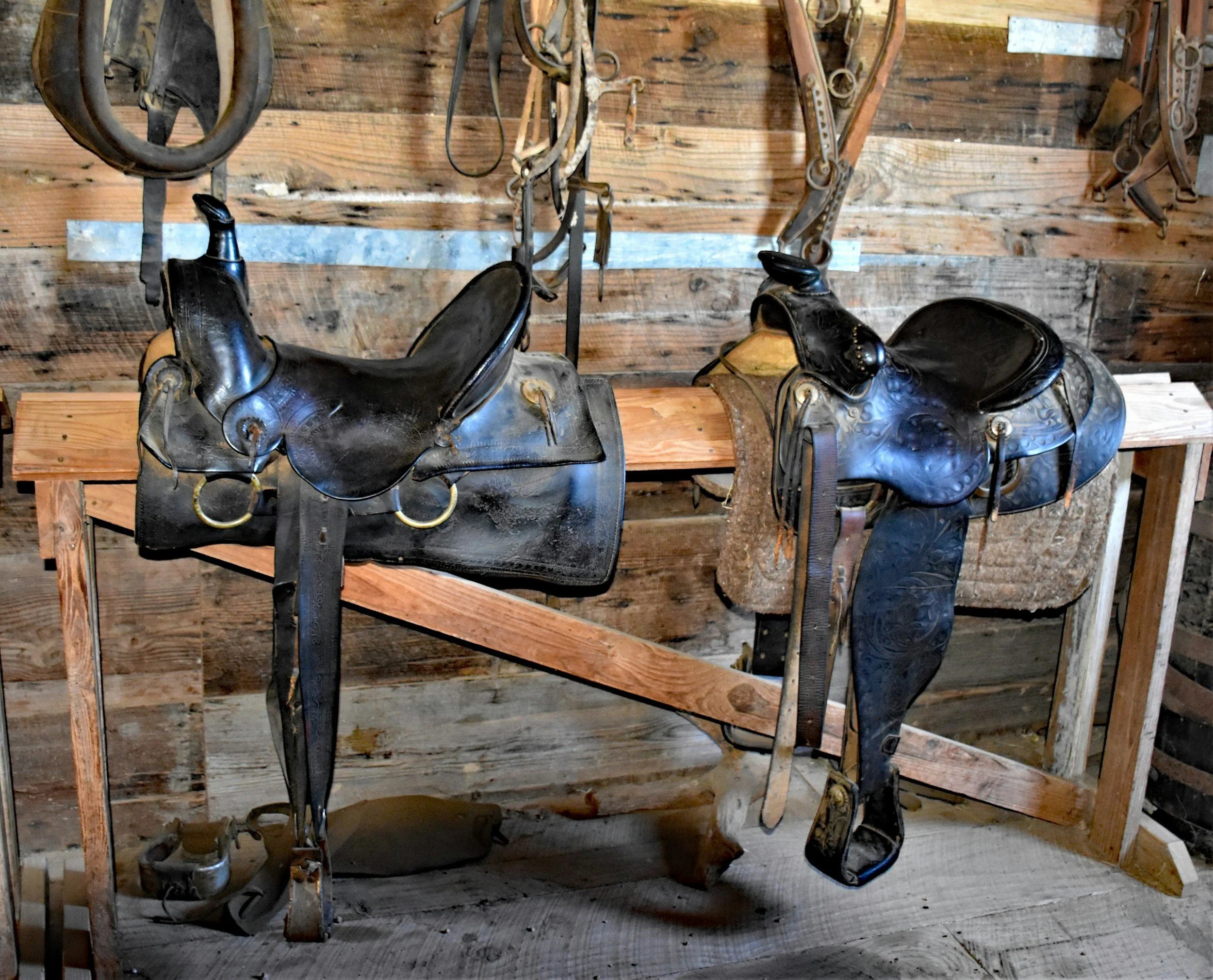 Two black leather Western horse saddles hanging on a wooden rack in a rustic barn.