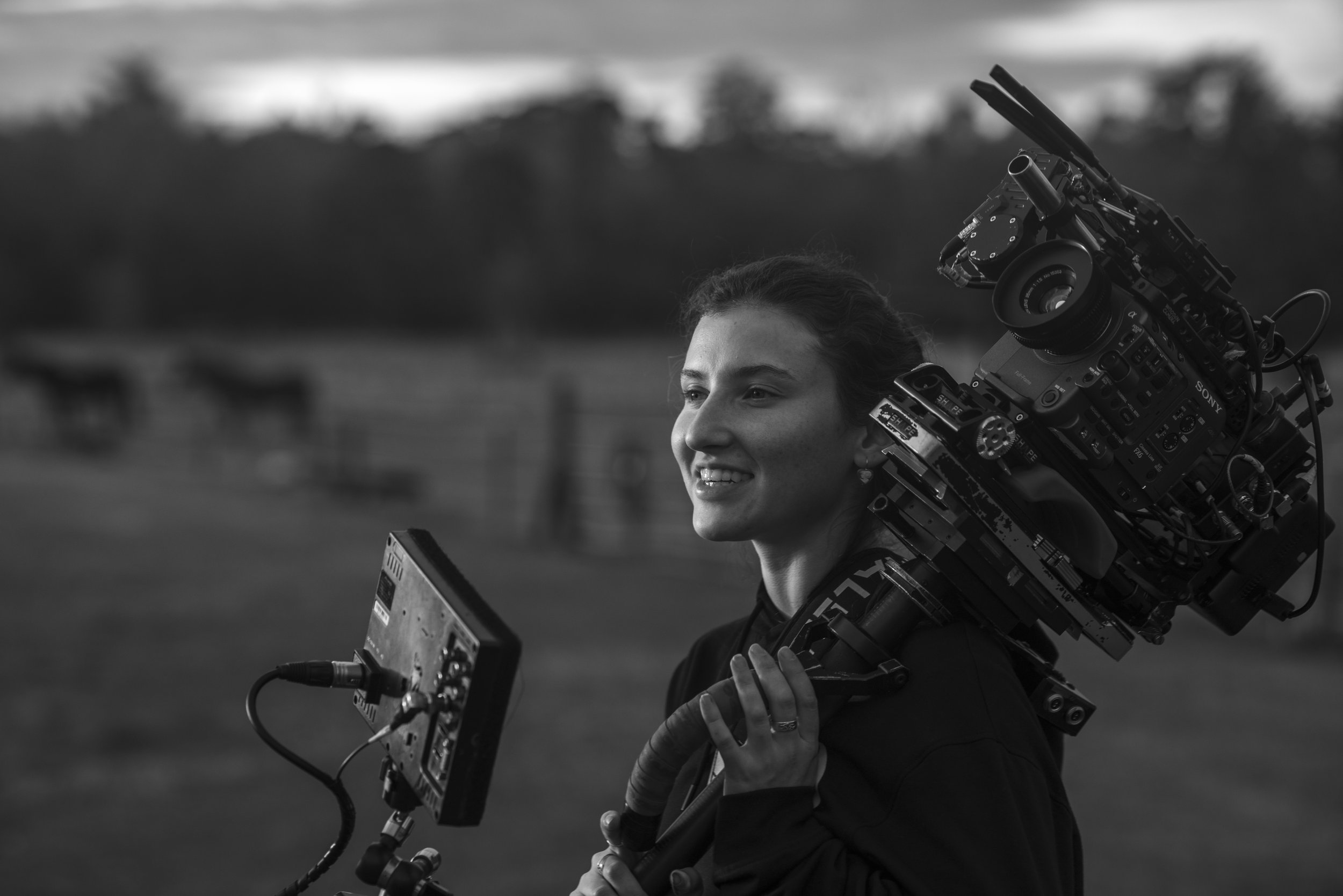 Molly McCormick from Kronos is carrying a professional film camera on her shoulder, holding a monitor in her hand, on the set of a music video.