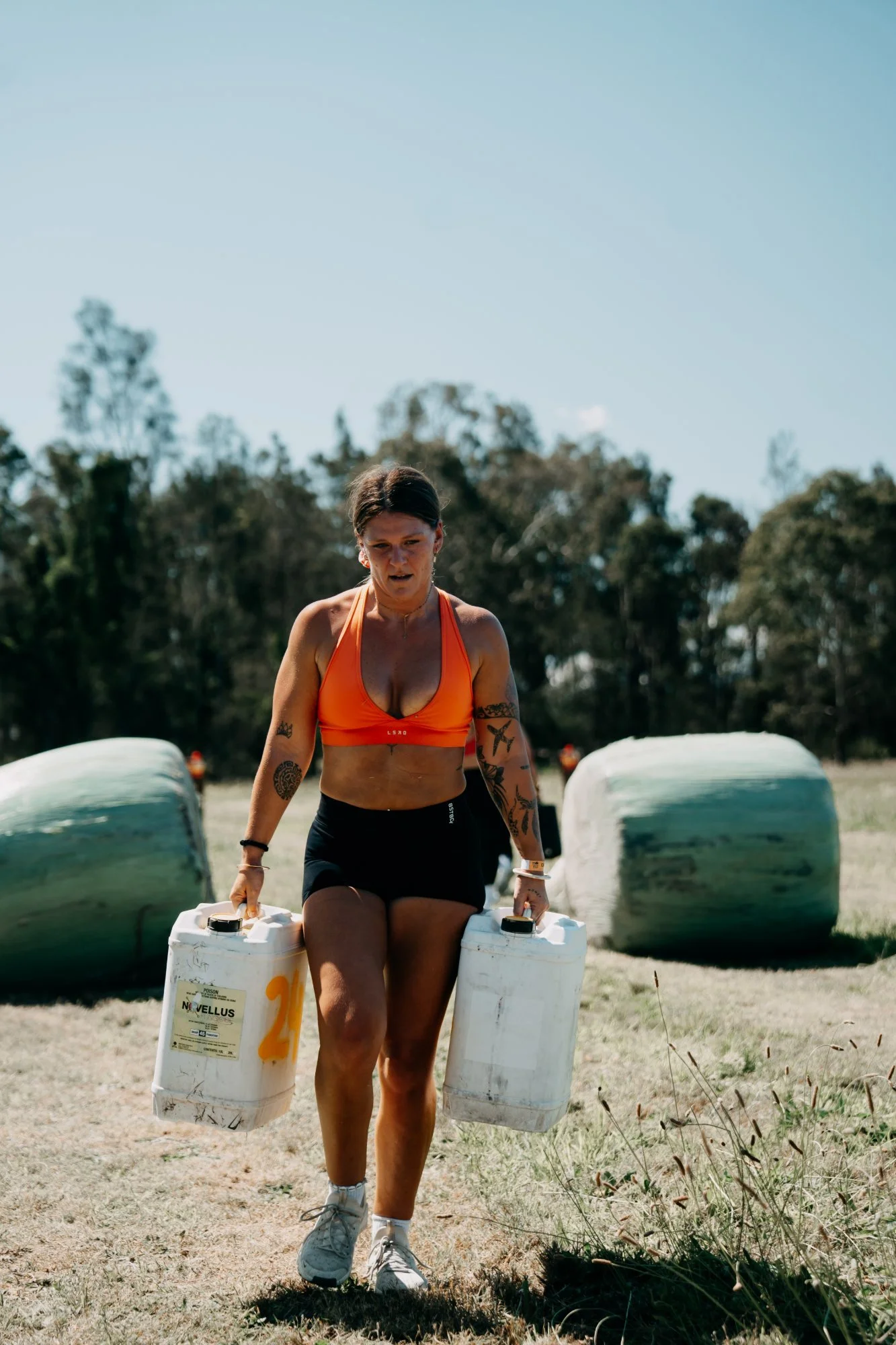Farm Strong competitor in an orange top doing a farmer's carry with two water drums past wrapped silage bales at The Farm Jam