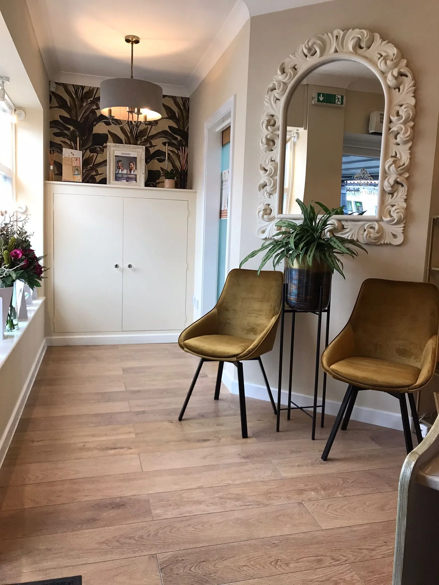 Interior of a waiting area with two mustard-colored chairs, a large green plant on a stand, a white decorative mirror, a white cabinet, and floral decorations on a window sill.