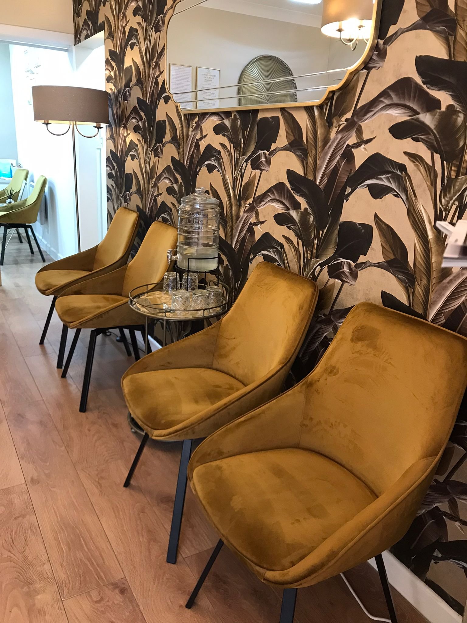 Three mustard yellow velvet armchairs are arranged along a wall with black and beige leaf-patterned wallpaper in a stylish indoor setting. A small side table with water dispenser and glasses is positioned next to the chairs. A mirror and wall-mounted