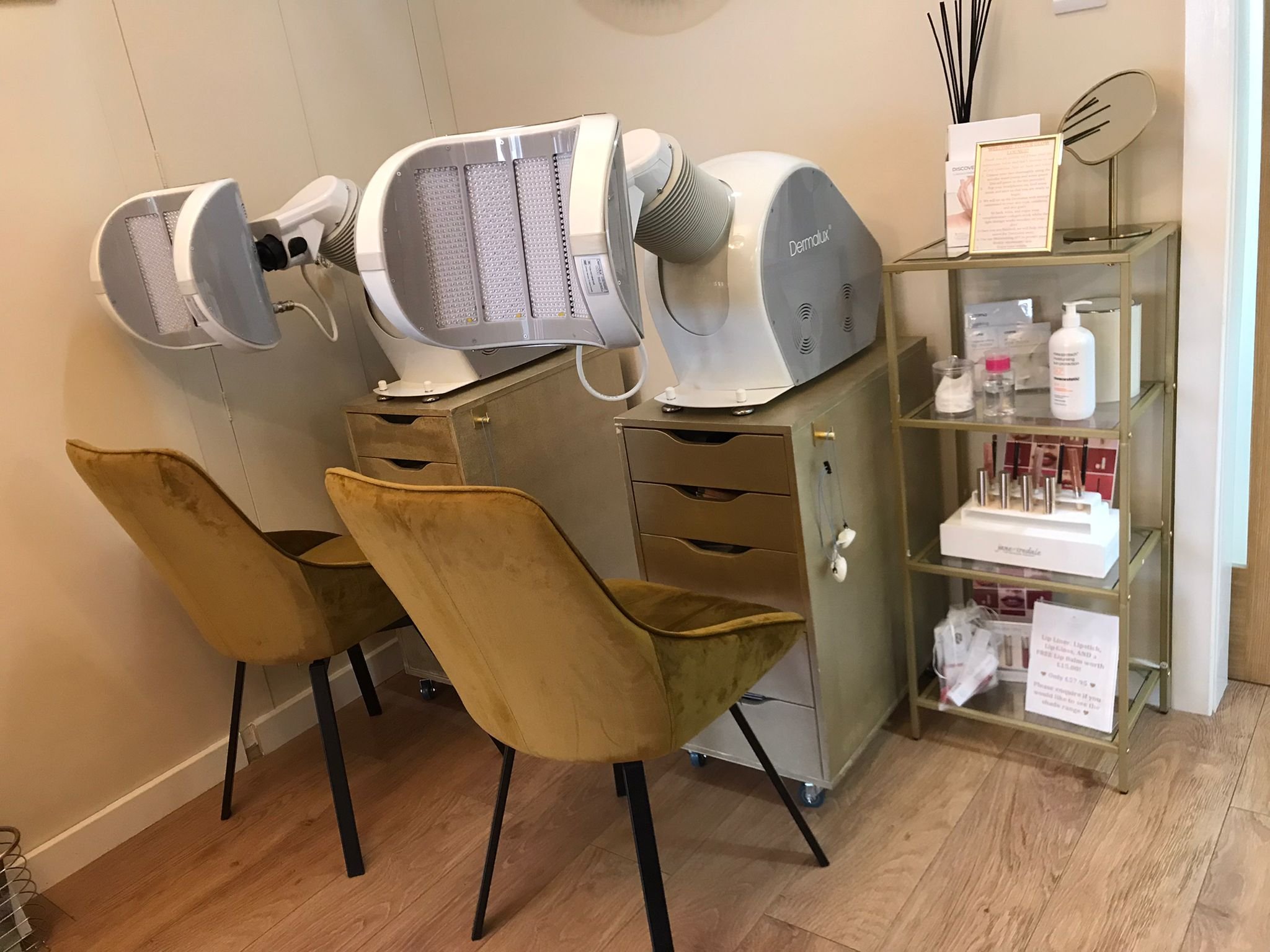 A skincare treatment room with two golden chairs, skincare devices on a counter, and a green shelving unit with skincare products and informational pamphlets.