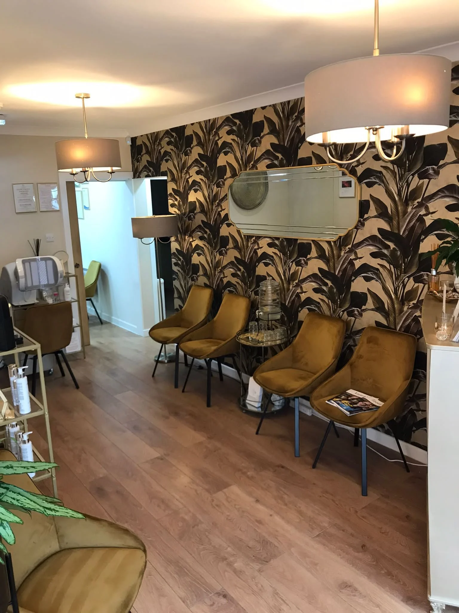 Interior of a waiting area with five mustard-colored chairs, a decorative mirror on a patterned brown leaf wallpaper, two ceiling lamps, a small side table with glasses and water bottle, and a partial view of a magazine.