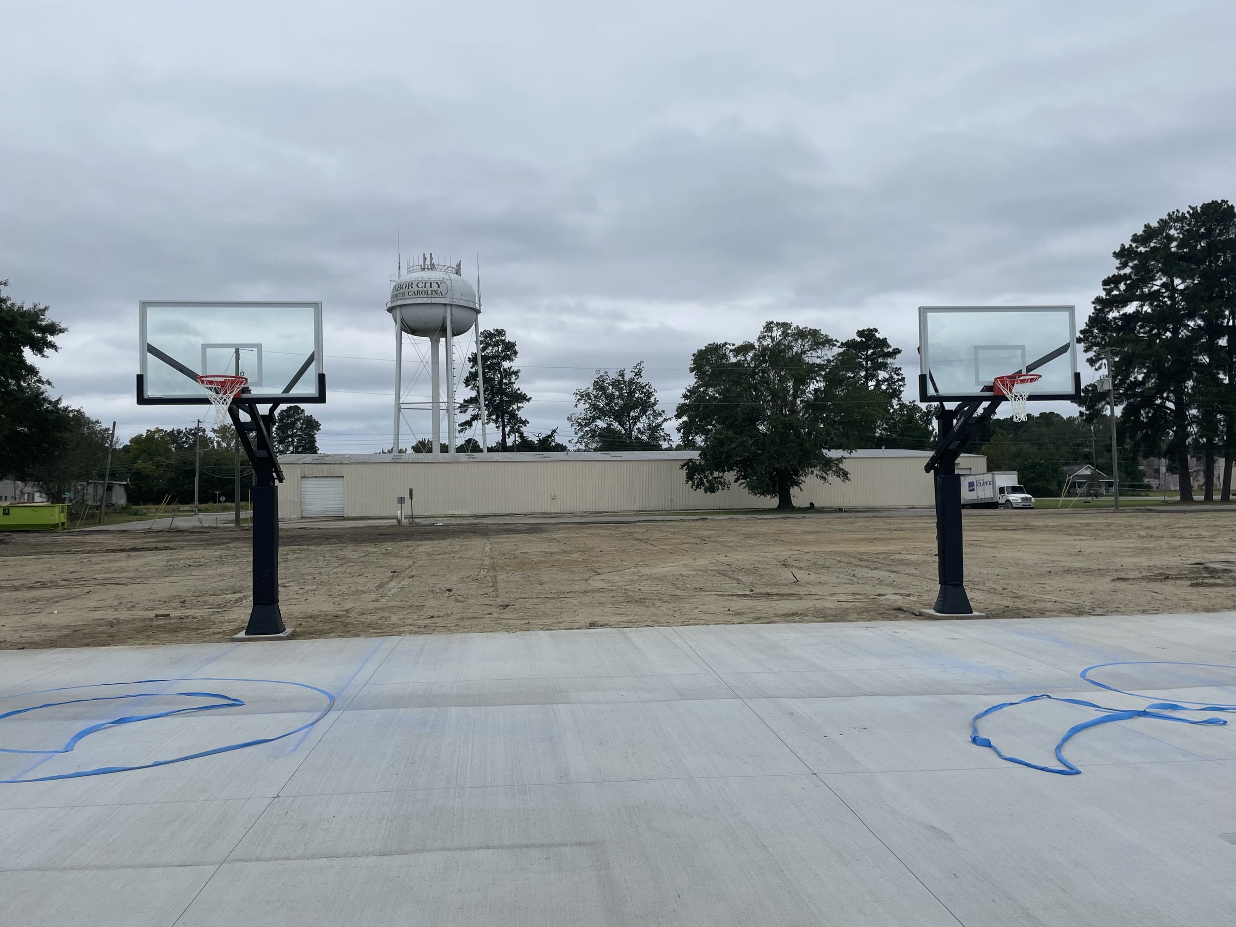 Outdoor basketball court with unfinished surface, featuring two basketball hoops and a water tower in the background, surrounded by trees and a building.
