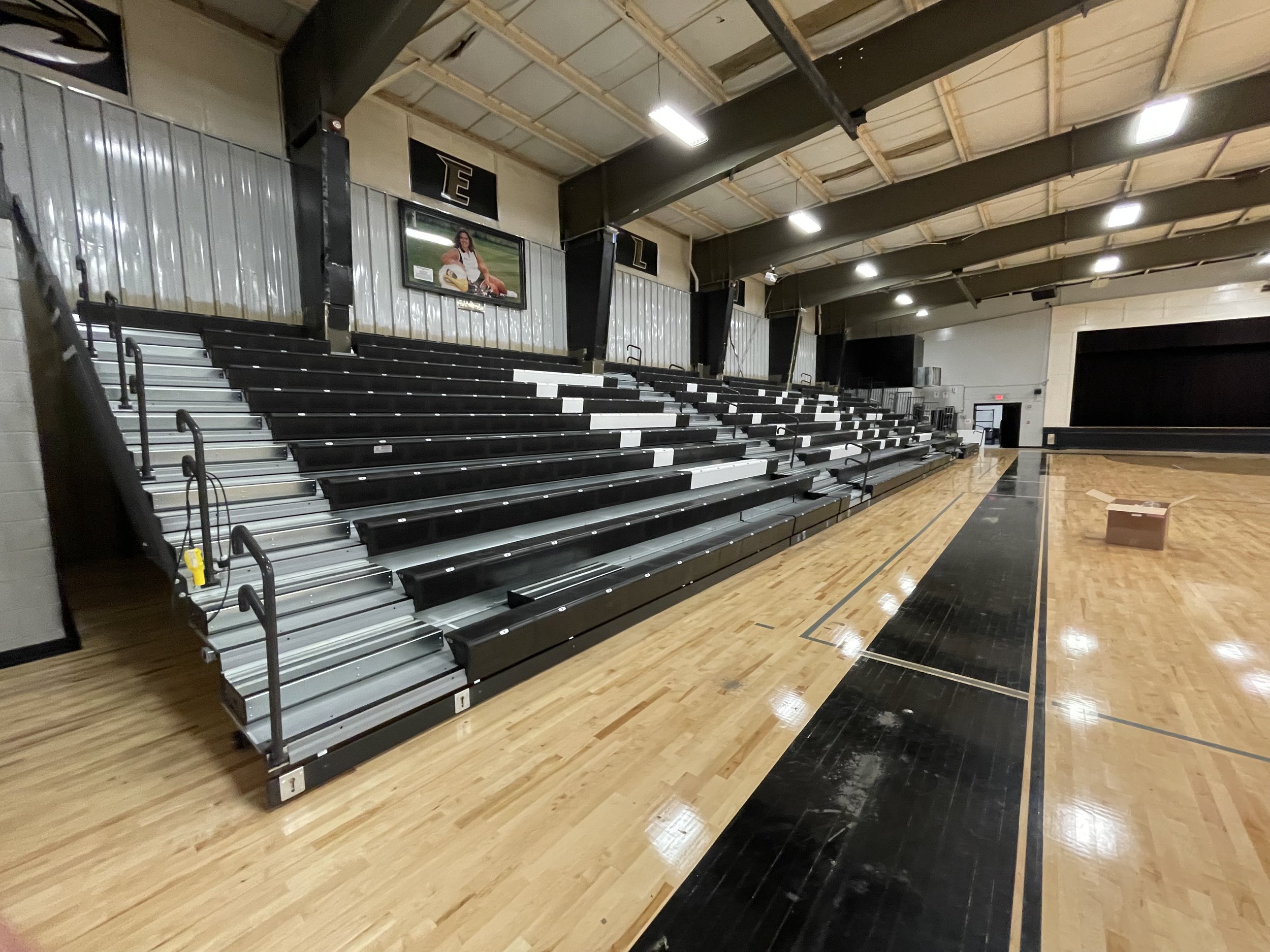 Indoor gymnasium with retractable bleachers, hardwood floor, and overhead lighting. A cardboard box is on the floor and there's a stage in the background.