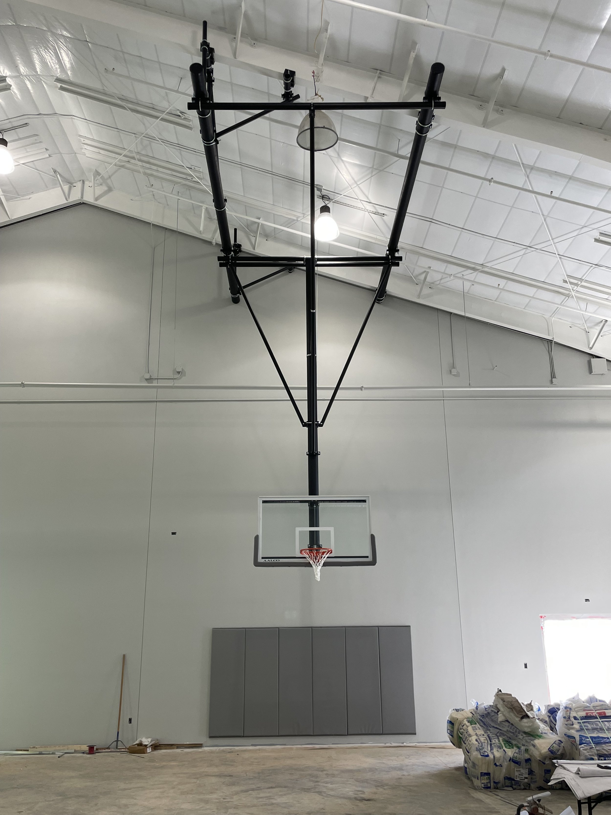 Indoor basketball hoop setup in a gymnasium under construction, with visible ceiling lights, wall padding, and construction materials on the floor.