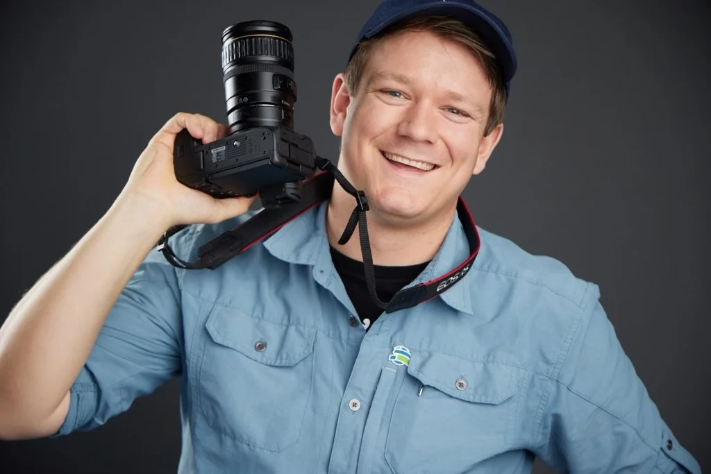 A smiling man holding a camera over his shoulder, wearing a blue shirt and cap, against a dark background.
