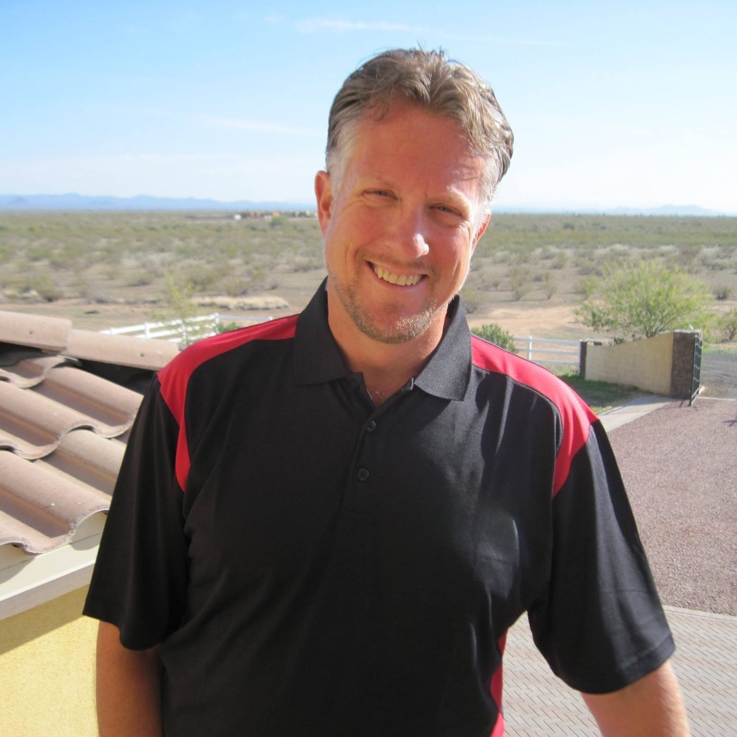 Smiling man outdoors in desert landscape wearing a black and red polo shirt.