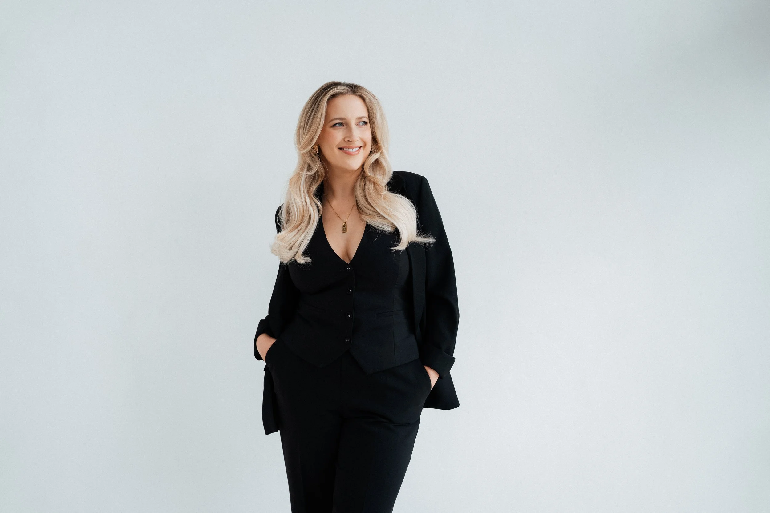 A woman with blonde hair wearing a black suit and necklace, standing against a plain light gray background, smiling and looking to the side.