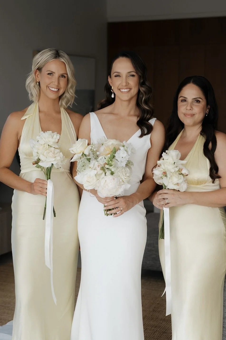 Three women in elegant dresses holding bouquets, standing indoors, likely at a wedding or formal event.