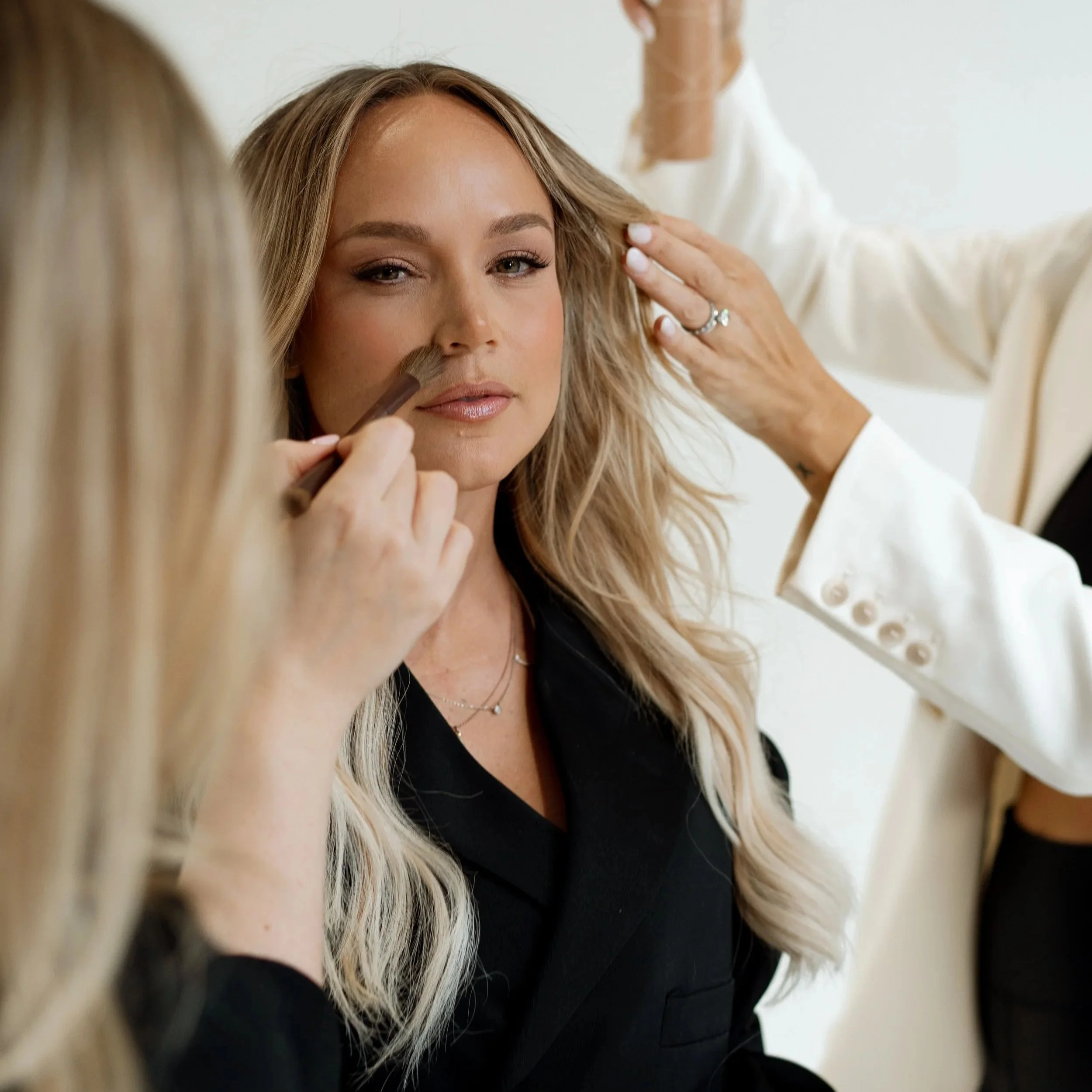 A woman having makeup applied by a makeup artist, with another person adjusting her hair in a white blazer.