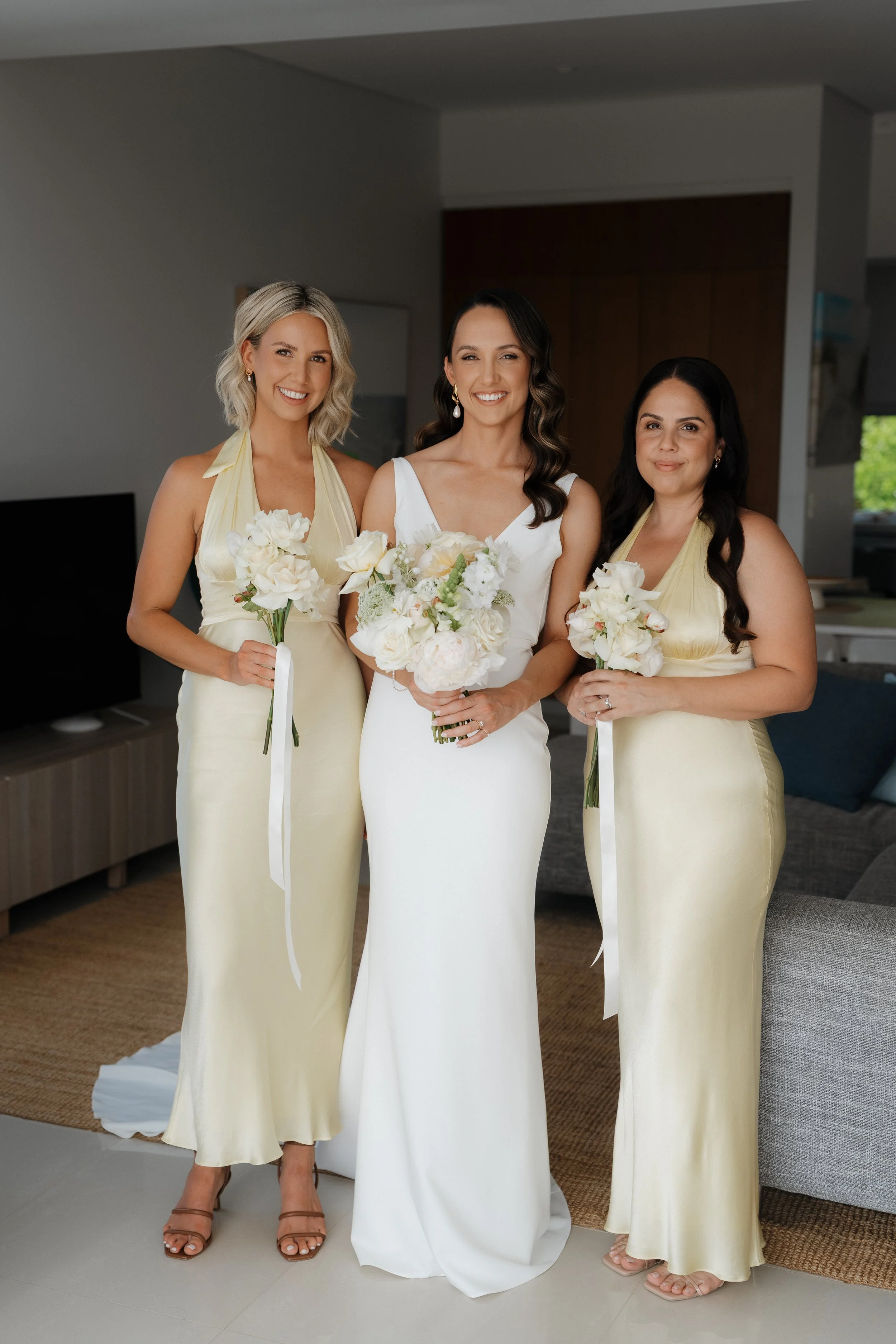 A bride and two bridesmaids standing indoors, smiling and holding bouquets of white flowers.