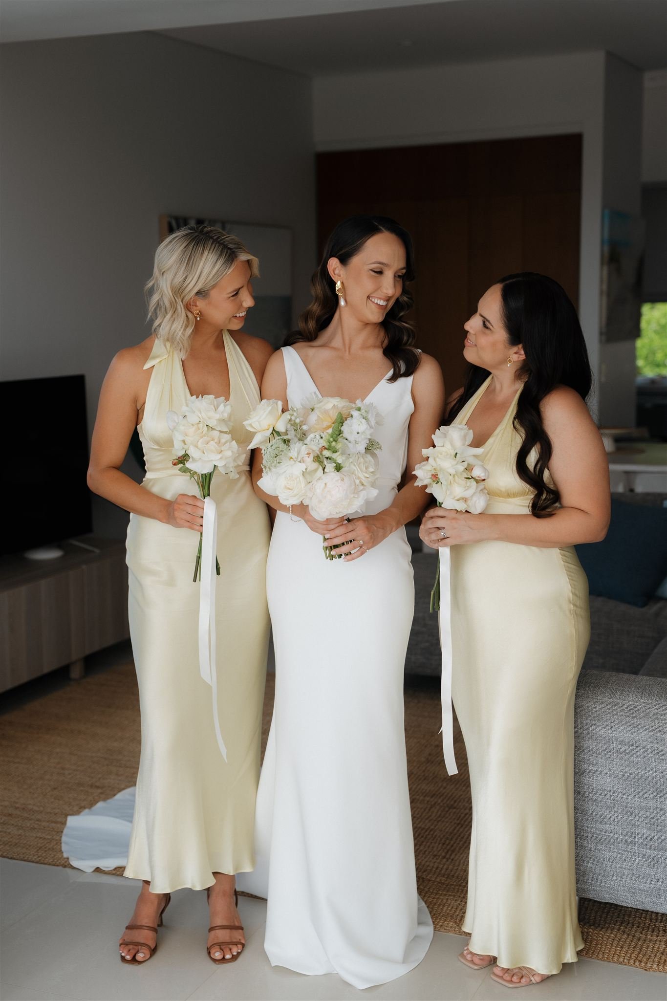 A bride in a white wedding gown holding a bouquet of white flowers, standing between two women in matching pale yellow dresses holding smaller bouquets, inside a modern room.