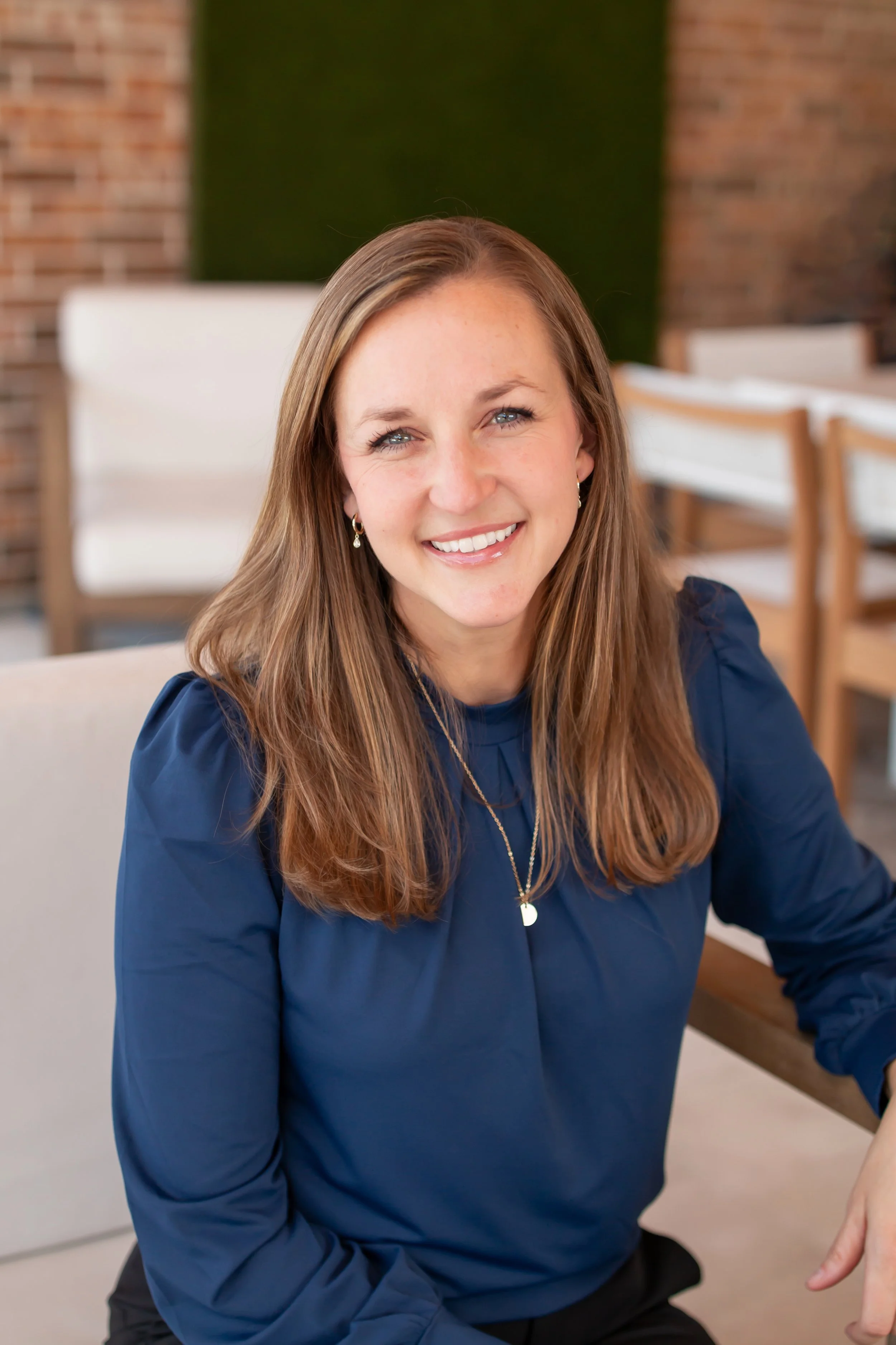 A woman with curly red hair sitting on a cream-colored sofa, smiling at the camera, wearing a light blue and white striped blouse.