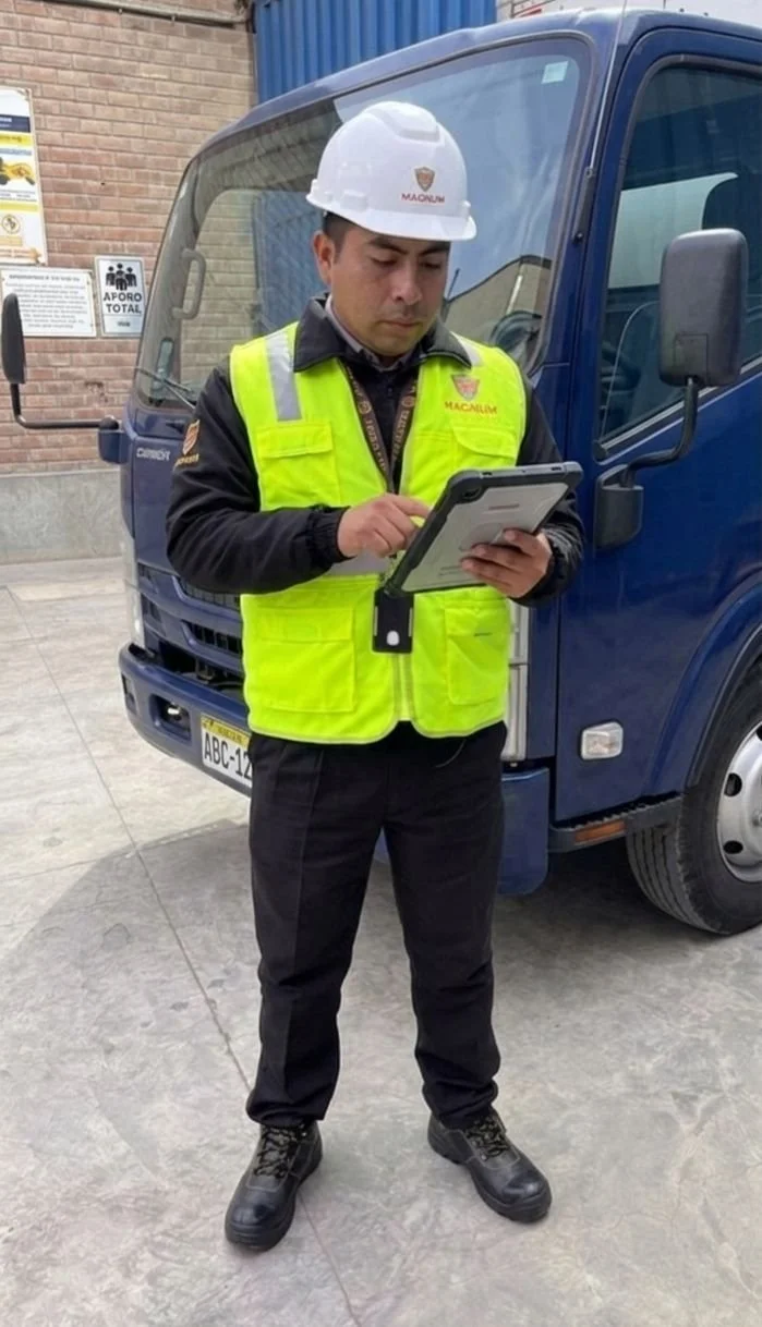 Hombre con casco y chaleco de seguridad revisando una tableta junto a un camión de color azul en un área industrial.