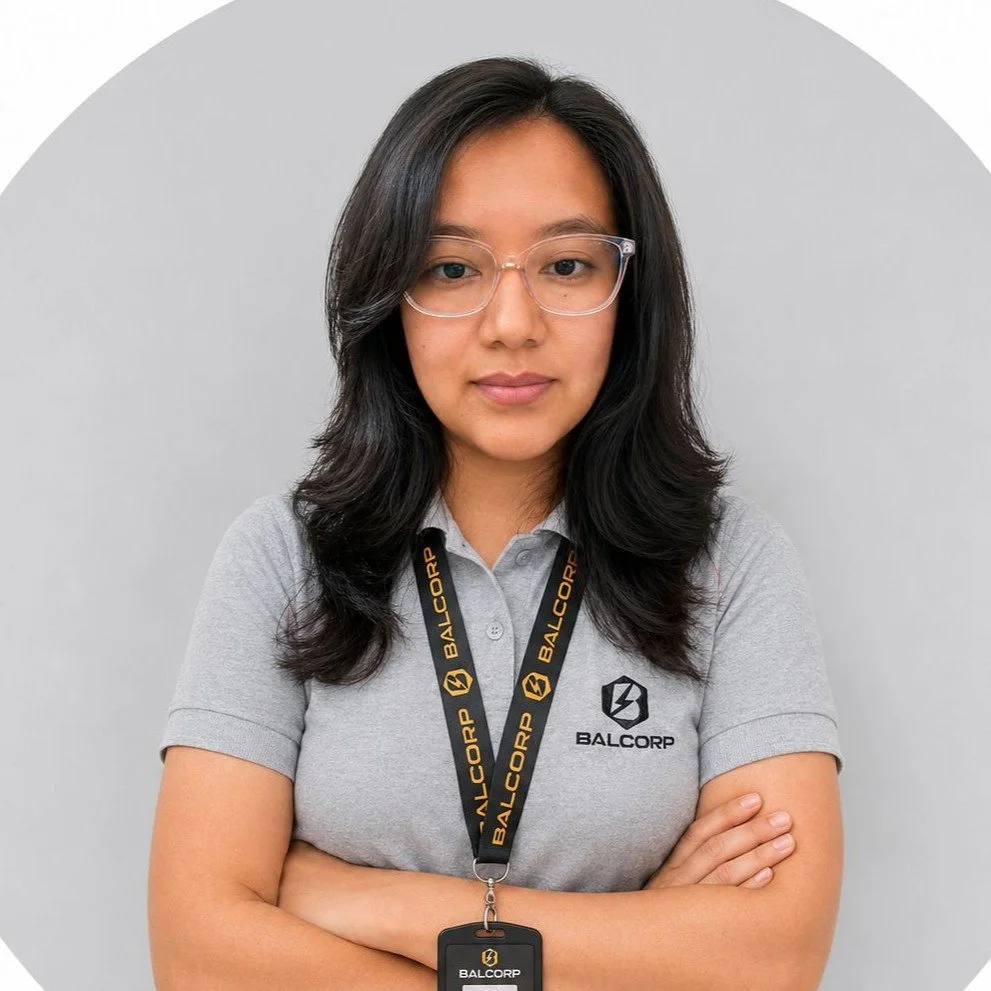 Una mujer con gafas y cabello largo, usando una camiseta gris con el logotipo de Balcorp y un gafete con la misma marca, de pie con los brazos cruzados frente a un fondo gris.