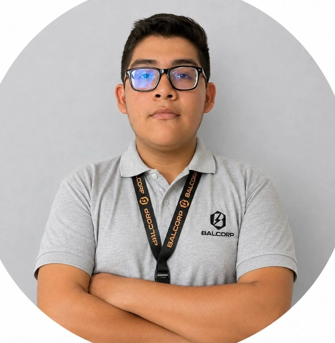 Joven con gafas, camiseta gris con logotipo de BALCORP, colgado de un collar con la marca BALCORP, brazos cruzados frente a una pared gris.