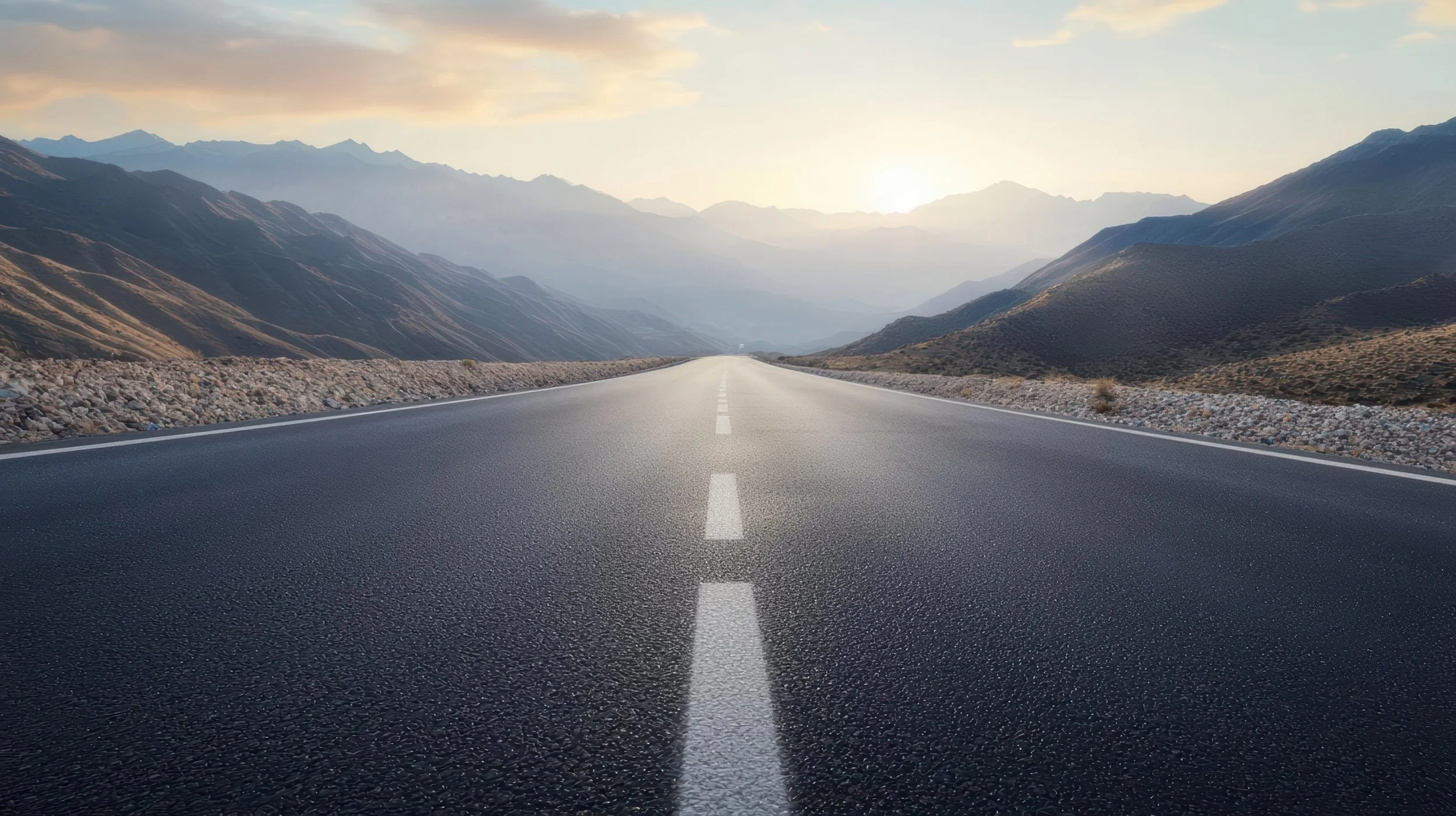 Empty straight asphalt road leading into the horizon between mountains with the sun rising or setting in the background.