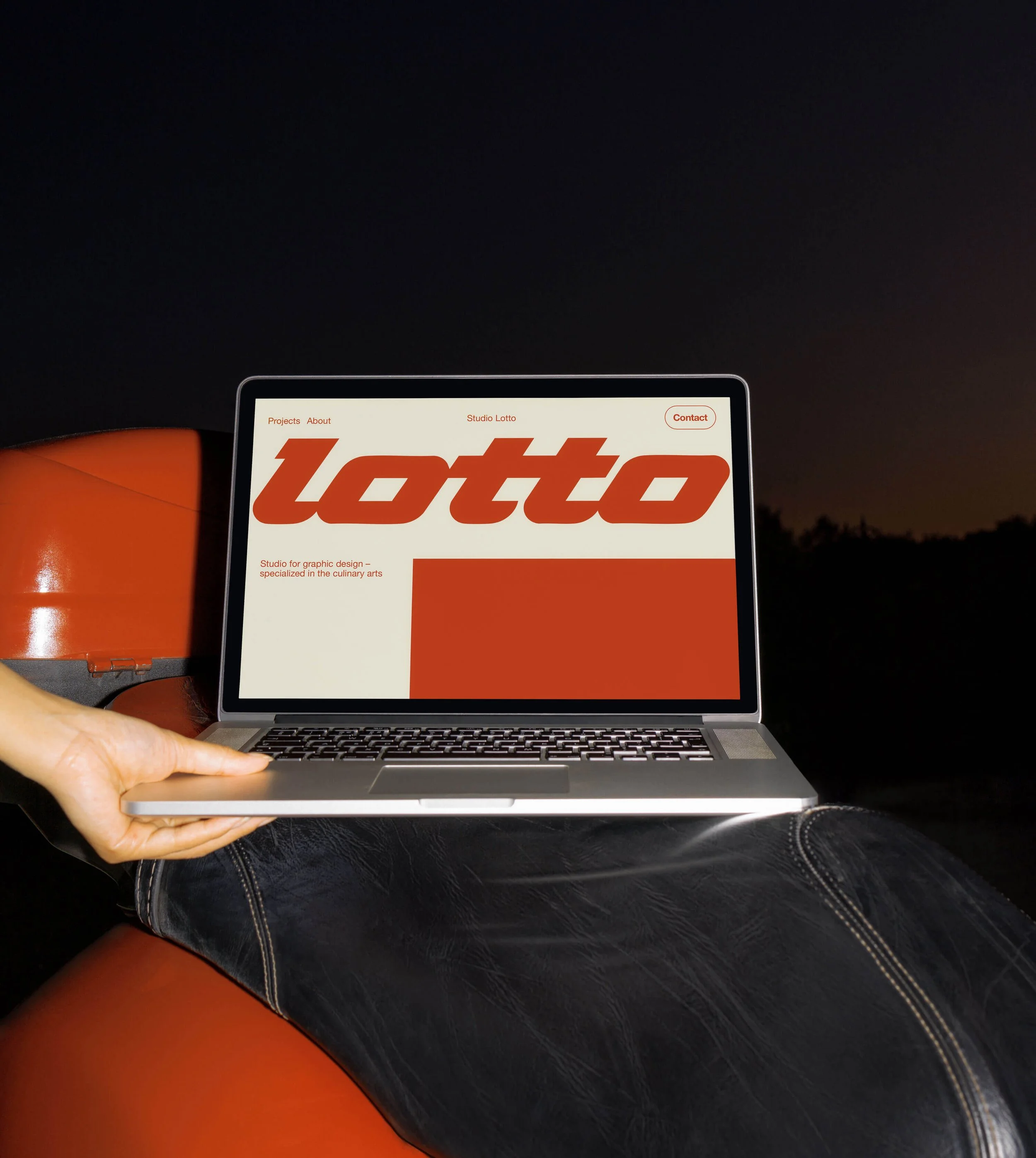 A person holding a silver laptop on their lap at night, displaying the website of Amsterdam-based Studio Lotto, with the word 'lotto' in bold red letters.