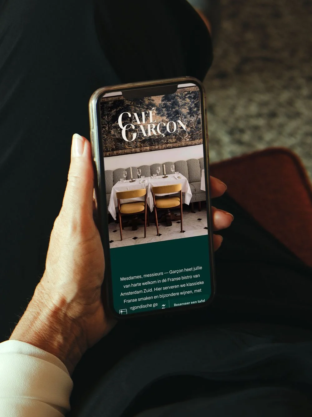 A person holding a smartphone displaying a website for Café Garçon in Amsterdam, featuring an image of a restaurant interior with a table set for dining and text in Dutch welcoming guests.