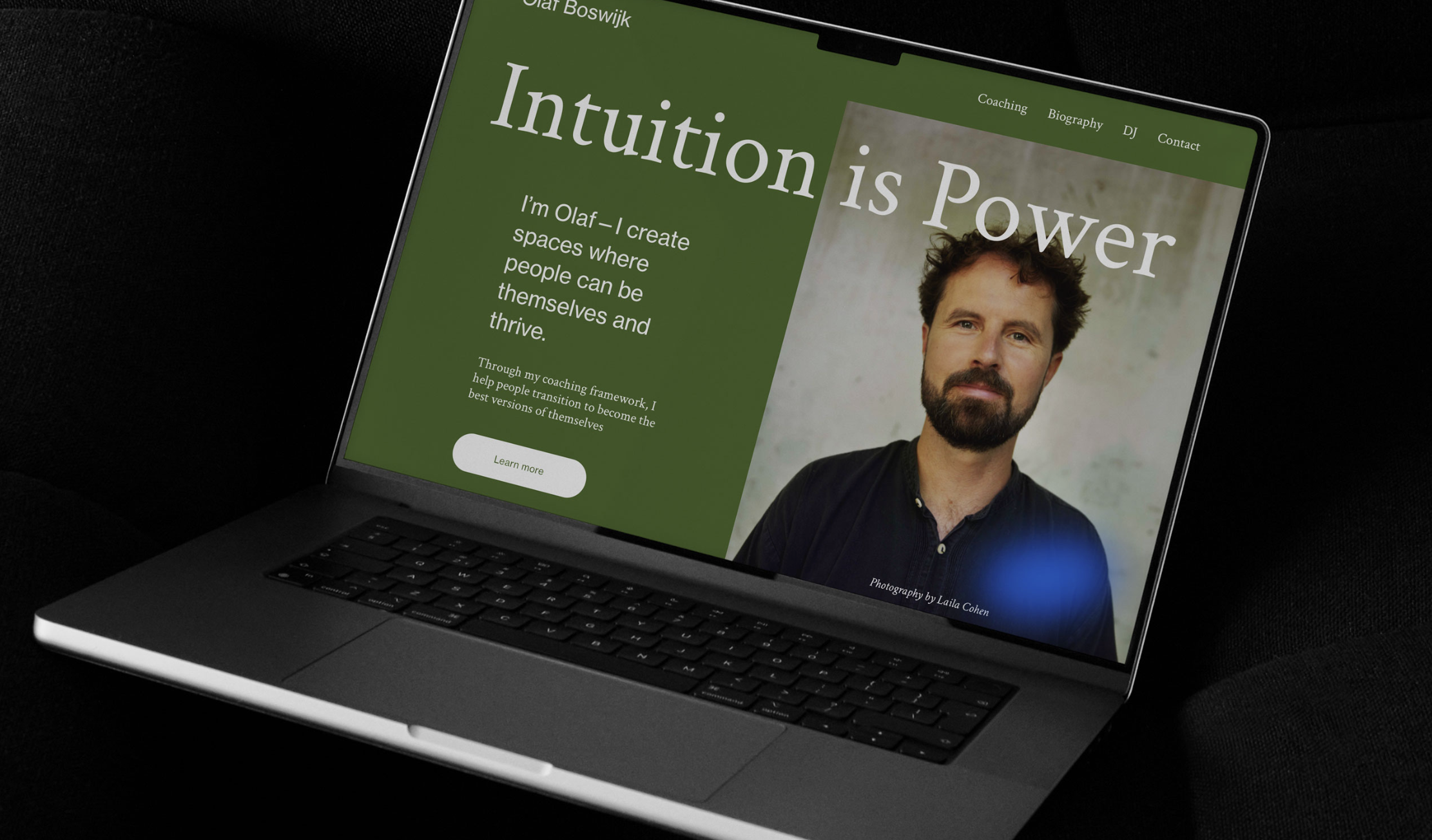 A MacBook laptop on a black surface displays a website about coaching and self-development. The website features a large photo of a man with brown hair and a beard, against a soft background. The page header reads 'Intuition is Power' with navigation