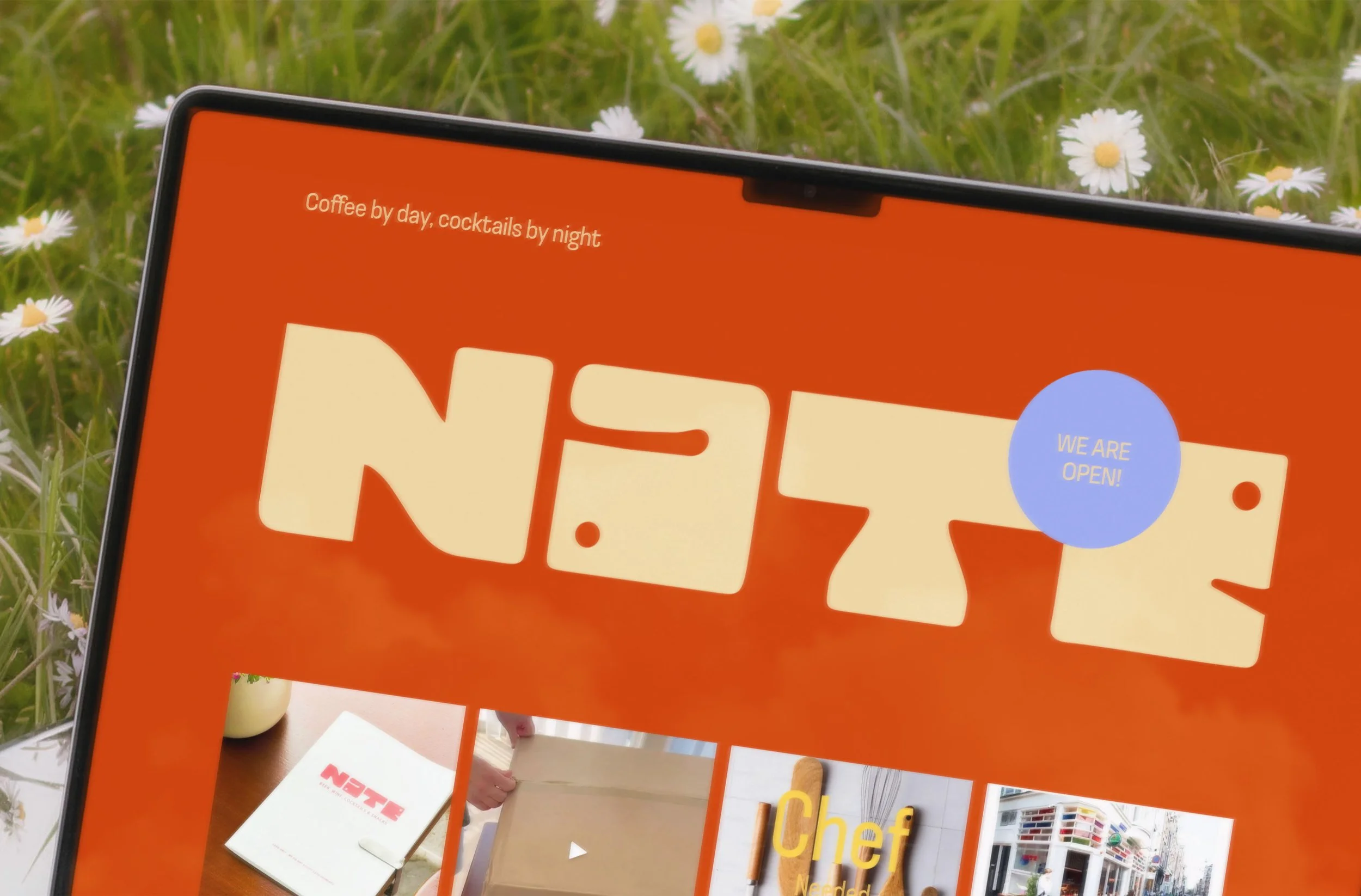Close-up of a tablet screen displaying the webpage of an Amsterdam-based restaurant called Nate. It has an orange background and a large yellow logo. The tablet is photographed outdoors on grass and daisies are in the background.