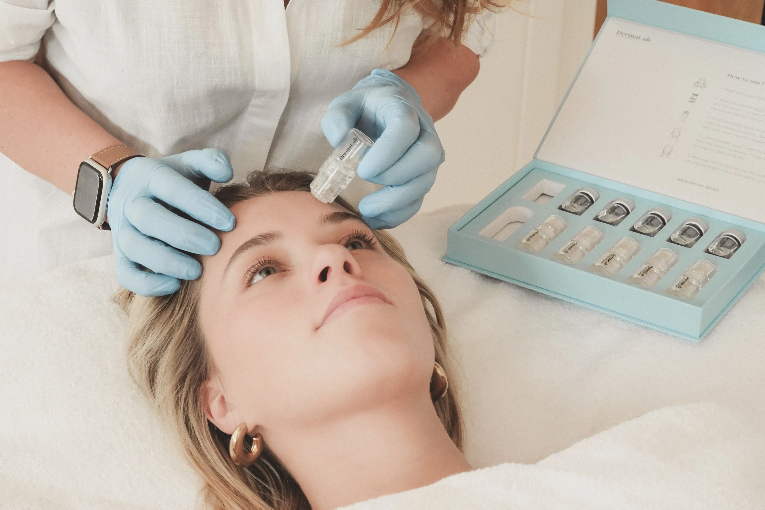 Een vrouw krijgt een huidbehandeling met een serum toegediend door een specialist in een kliniek, met een set met ampullen op de tafel ernaast.