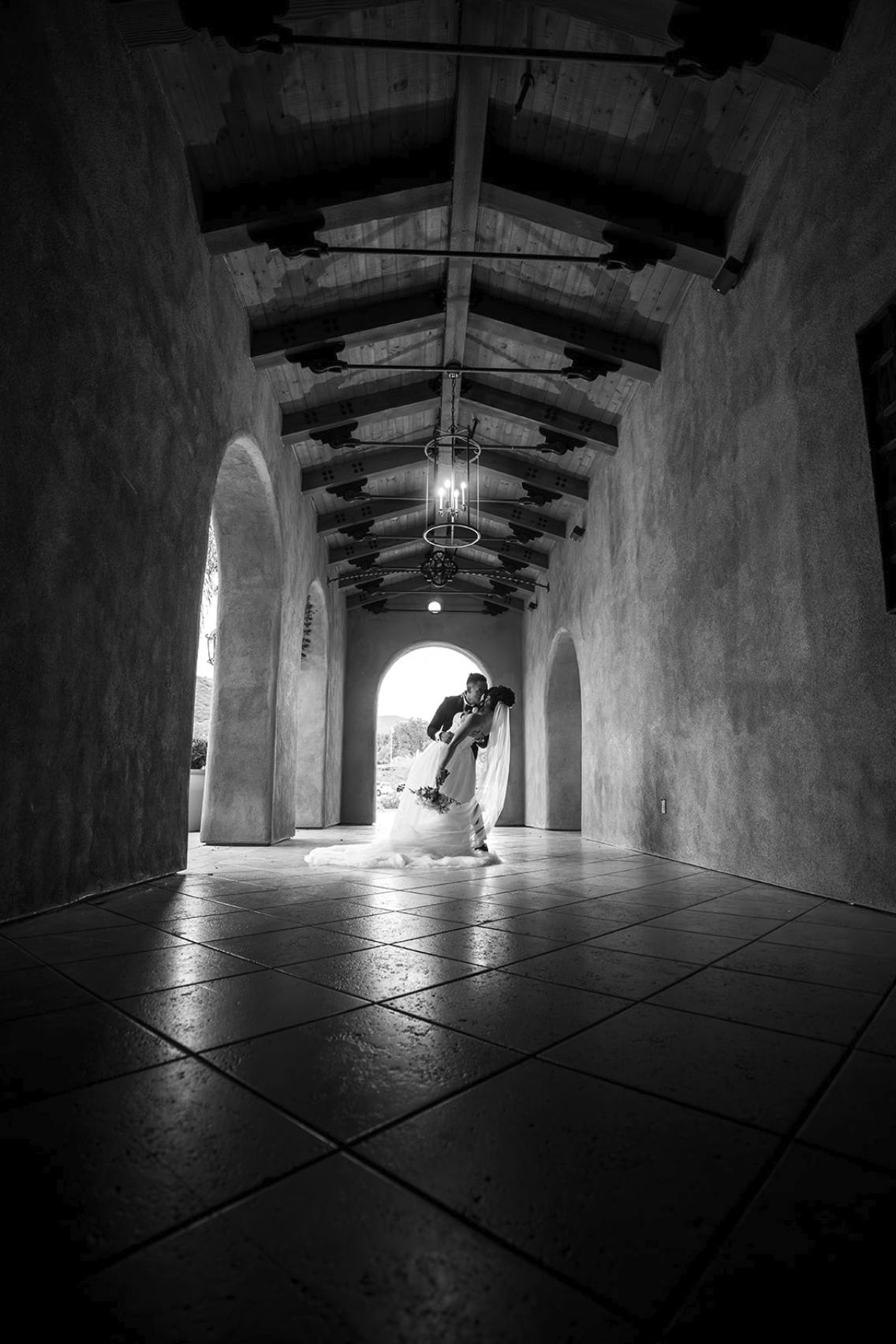 A black and white photo of a bride and groom sharing a kiss in an arched hallway with tiled floor, exposed wooden beams, and hanging chandelier.