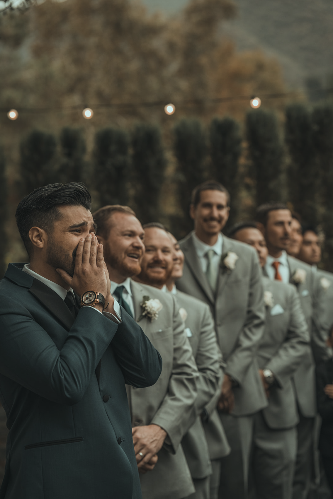 A man in a dark suit is crying with his hands covering his face, surrounded by other men in light gray suits at an outdoor wedding.