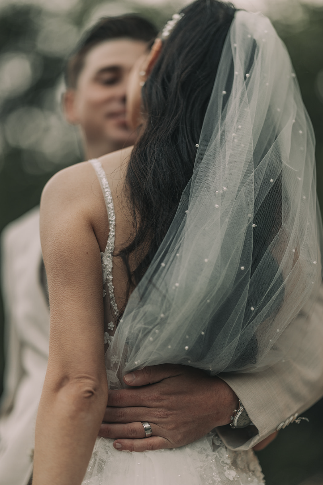 A bride and groom embracing outdoors during their wedding, with the bride wearing a veil and wedding dress, and the groom in a light-colored suit.