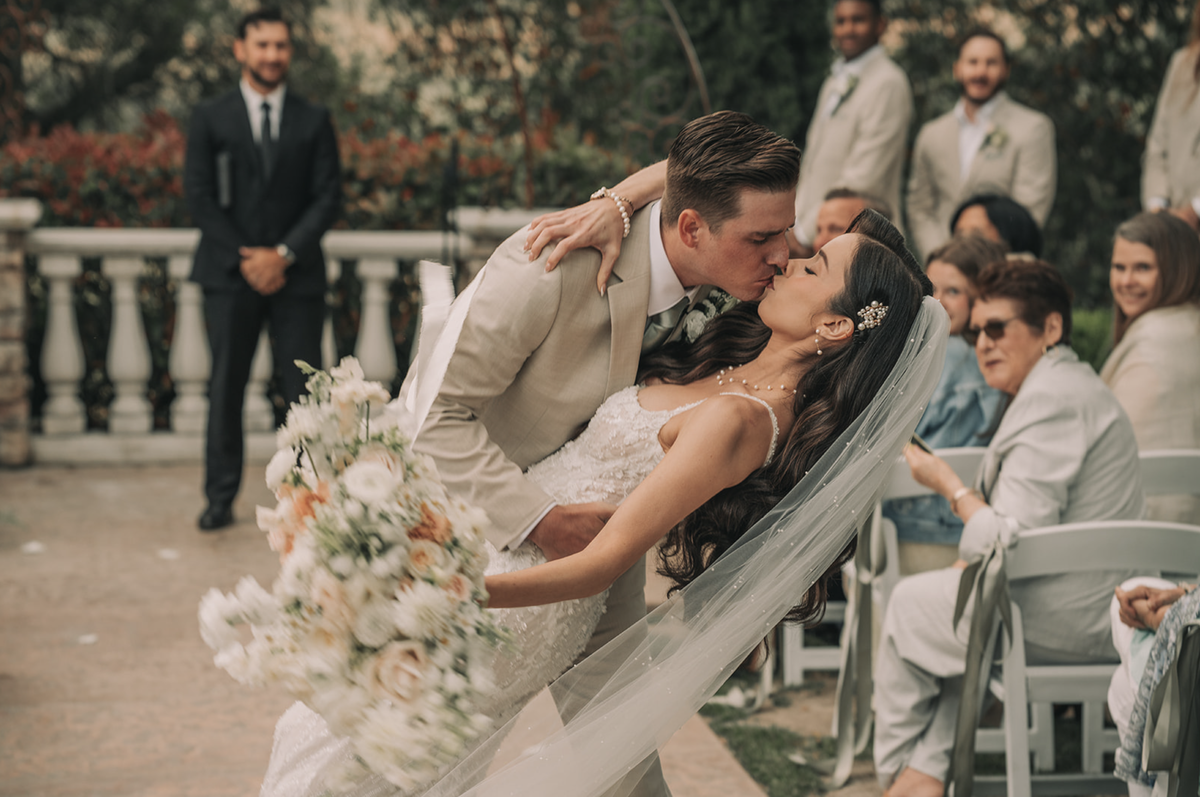 A newlywed couple sharing a kiss during their outdoor wedding ceremony, with guests seated nearby and the groom dipping the bride backwards. Wedgewood wedding venue Chino Hills.