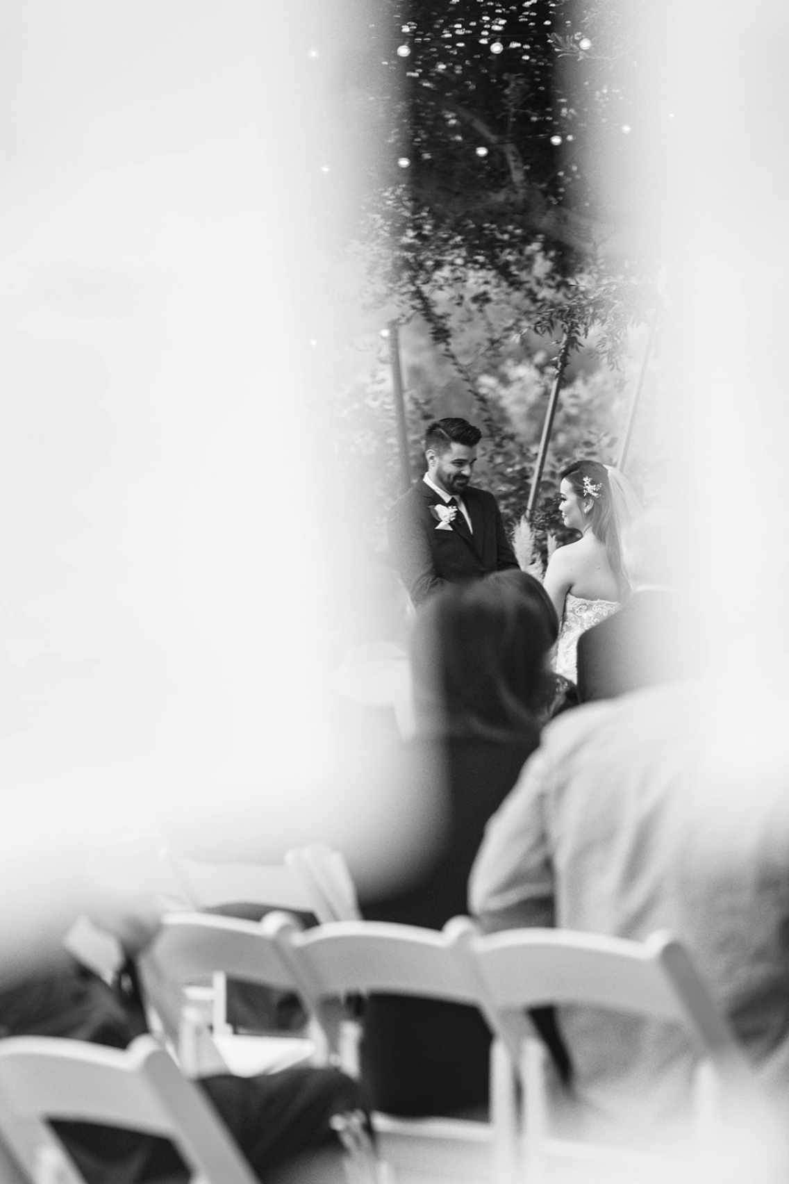 Black-and-white photograph of a wedding ceremony, with a groom and bride facing each other under a leafy outdoor setting. The groom is wearing a dark suit with a boutonniere, and the bride is in a strapless wedding gown with floral embellishments in 