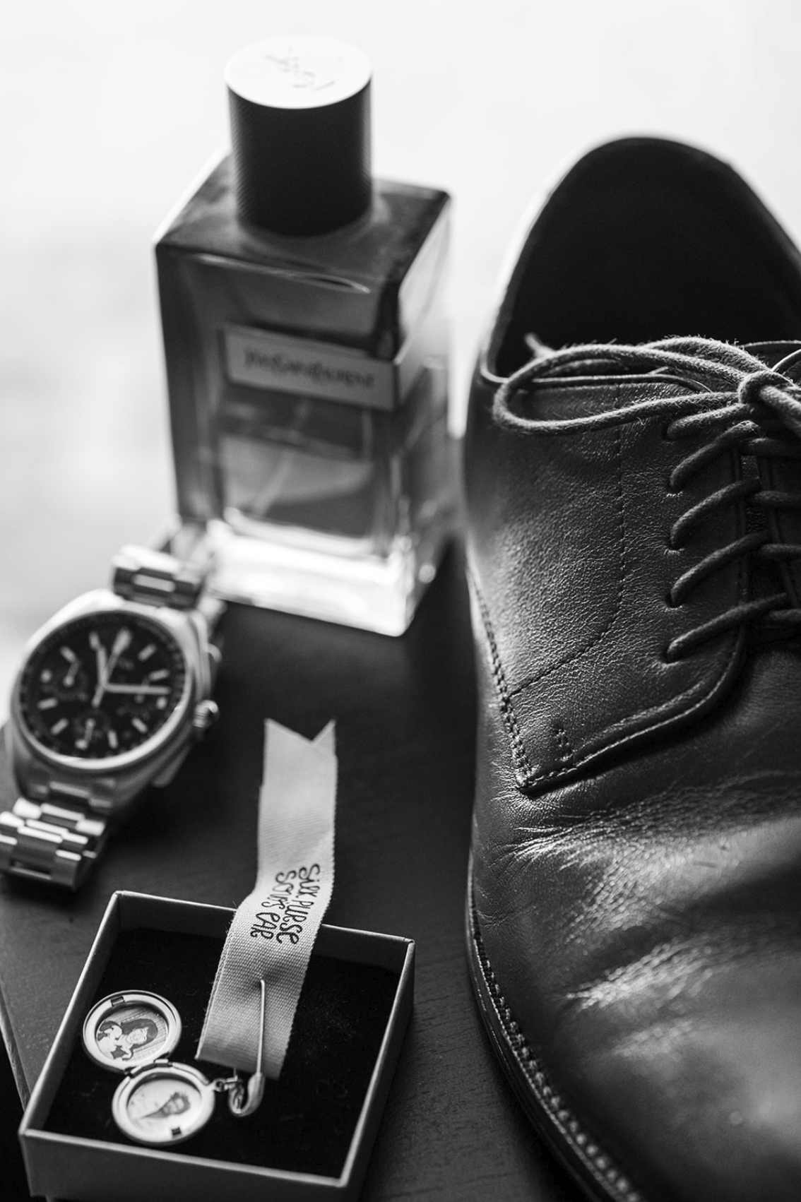 Black leather shoe, wristwatch, perfume bottle, and a box of cufflinks on a dark surface.
