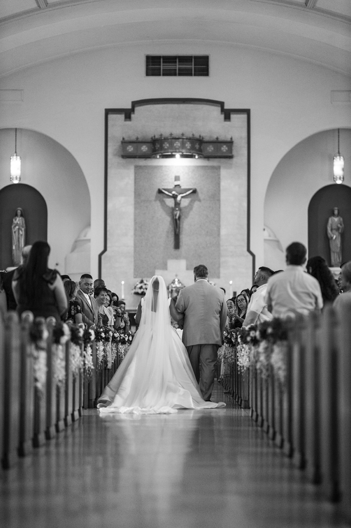 A bride and a man walking down the aisle of a church during a wedding ceremony. The church interior features religious statues and a crucifix at the altar.