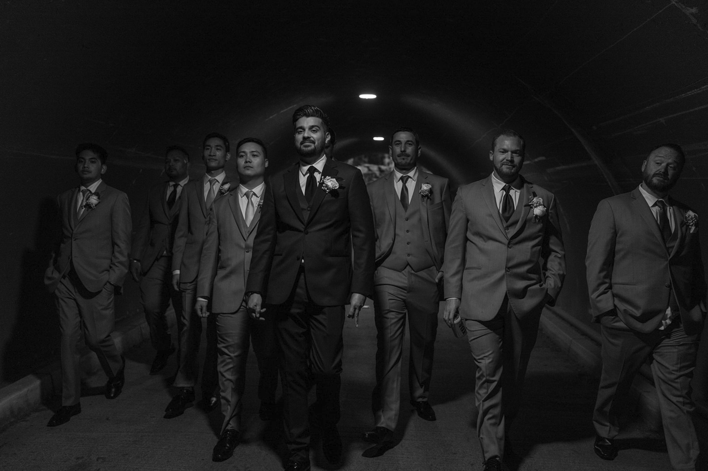 Black and white photo of a groom and seven groomsmen walking through a tunnel, dressed in suits with boutonnières.
