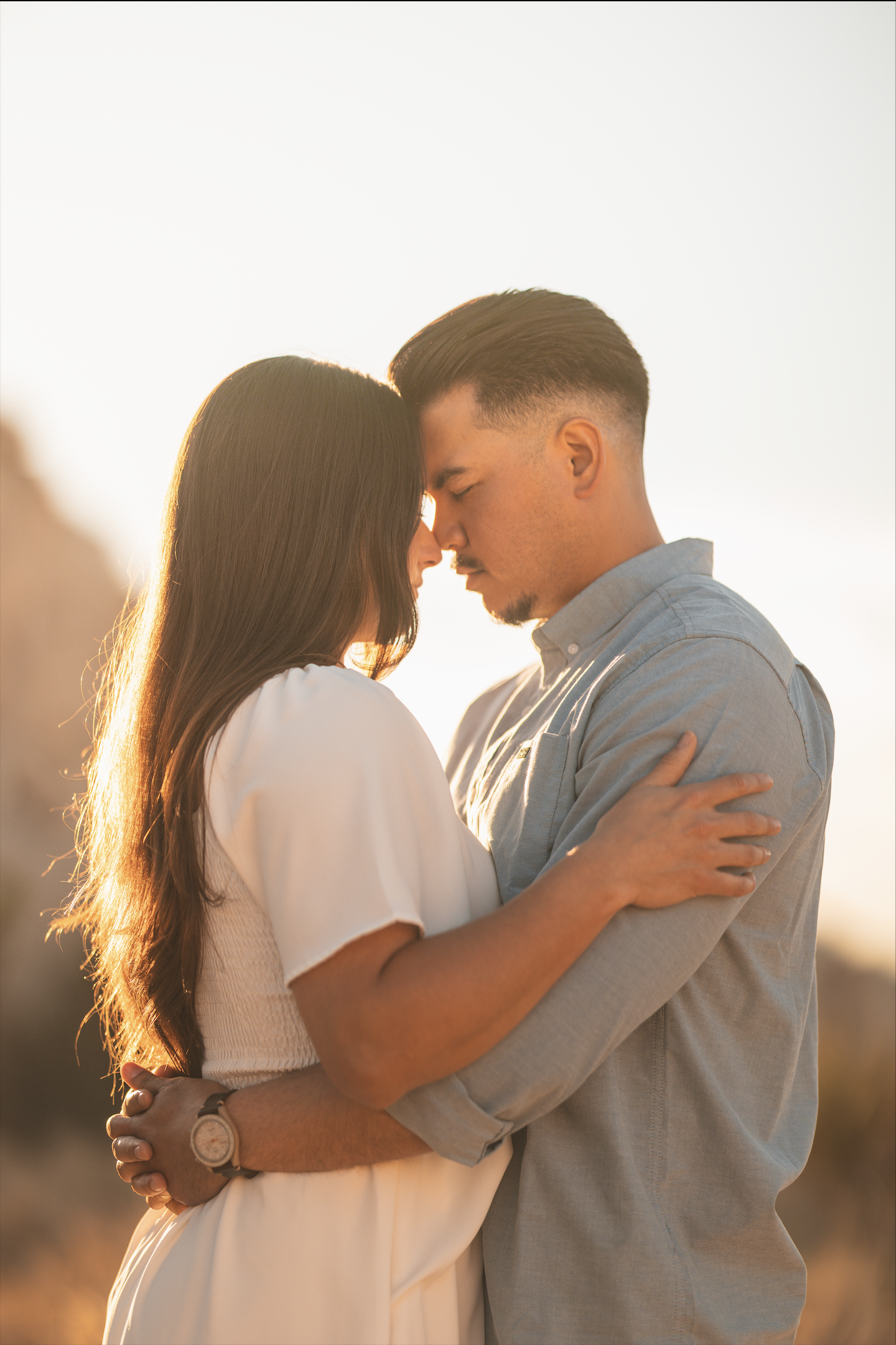 A romantic couple with their foreheads touching, holding each other close outdoors during sunset.