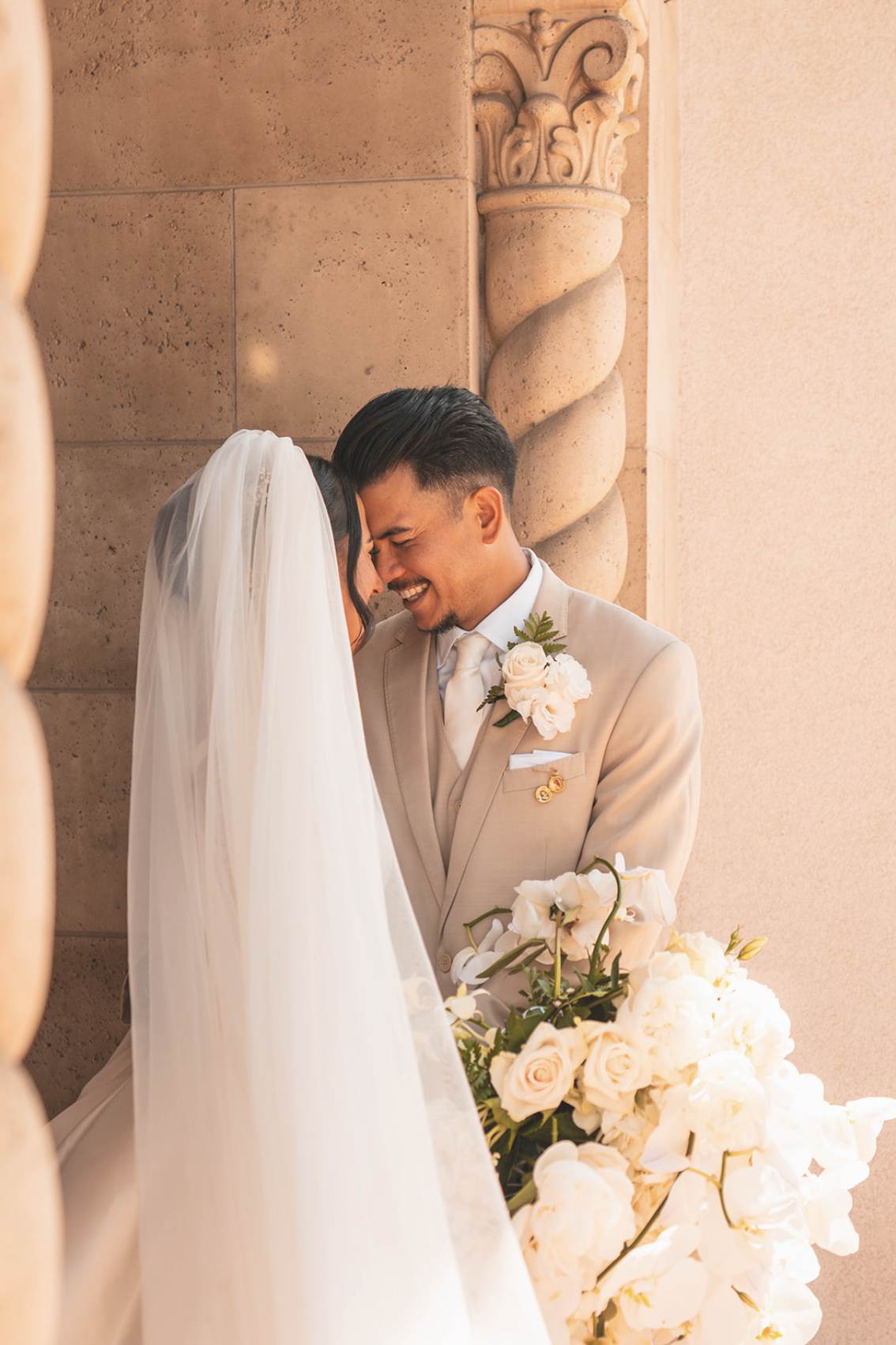 A bride and groom share a tender moment at their wedding, touching foreheads and smiling, with the bride wearing a white veil and holding a large bouquet of white flowers. The groom is dressed in a beige suit with a white shirt and tie, adorned with 