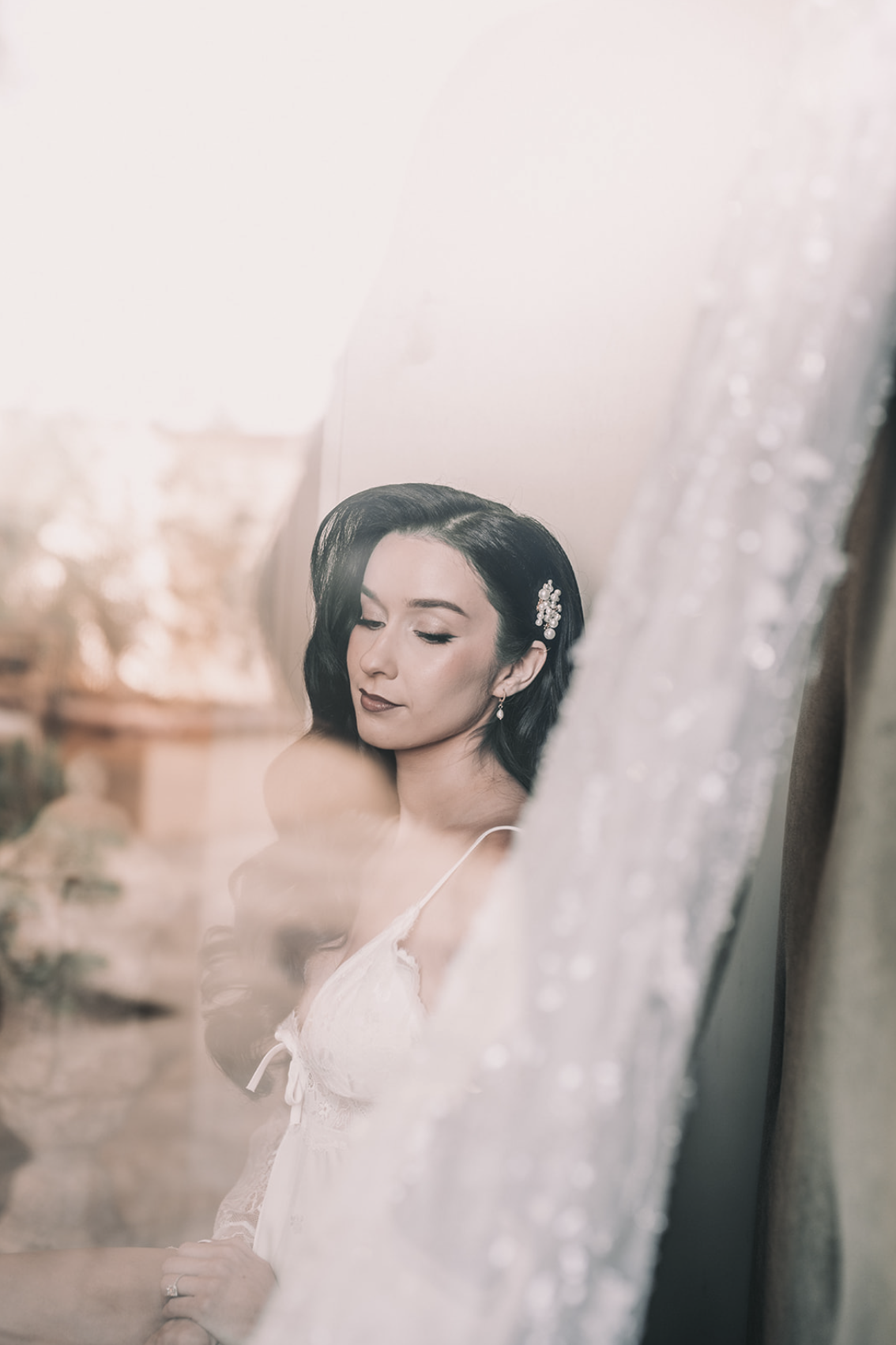 A woman with black hair styled in vintage waves, wearing earrings and a spiked hair accessory, seen through a window with a veil decorated with beads, looking down with closed eyes.