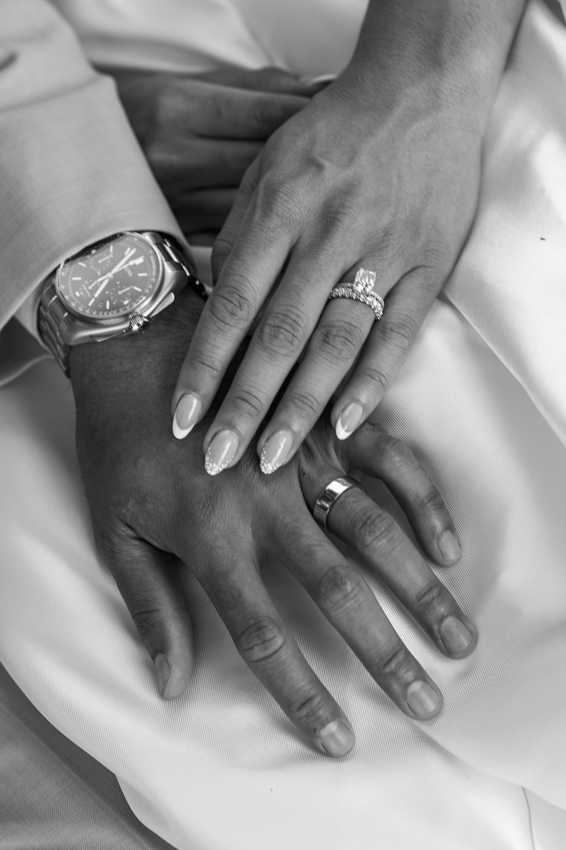 Close-up of a couple's hands, with wedding rings, resting on a white surface. The man wears a watch, and the woman has manicured nails with decorative tips. Black and white photo.