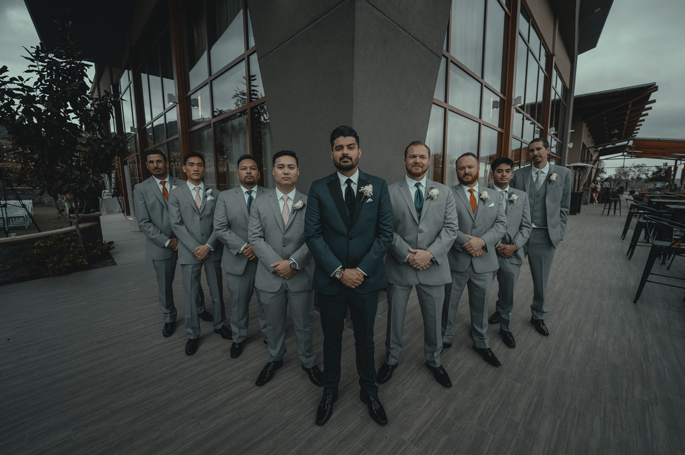 A group of nine men dressed in suits standing outside a modern building with large glass windows and wooden accents, looking at the camera.