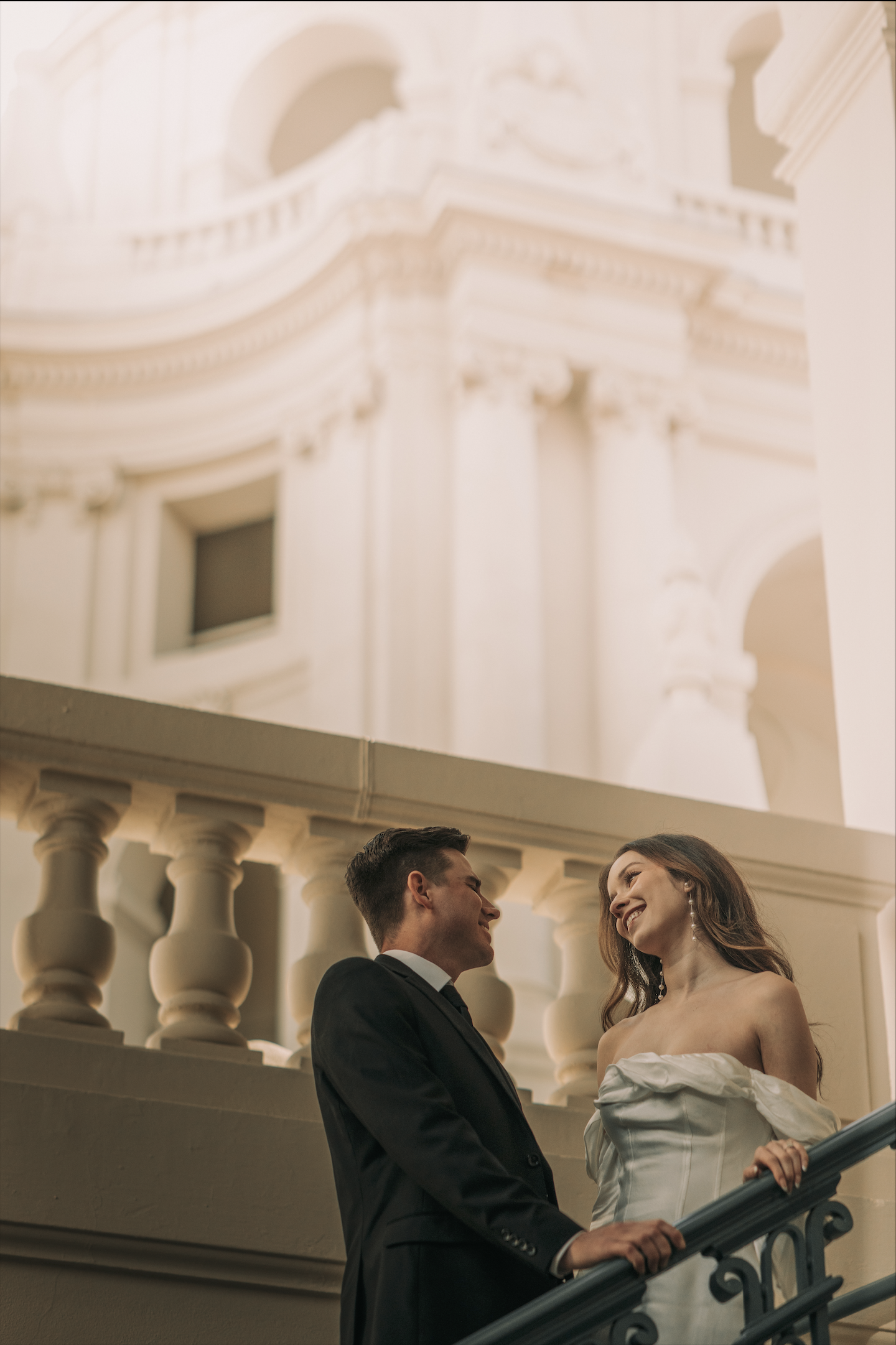 A couple dressed in formal attire sharing a happy moment on a staircase inside a grand, ornate building.