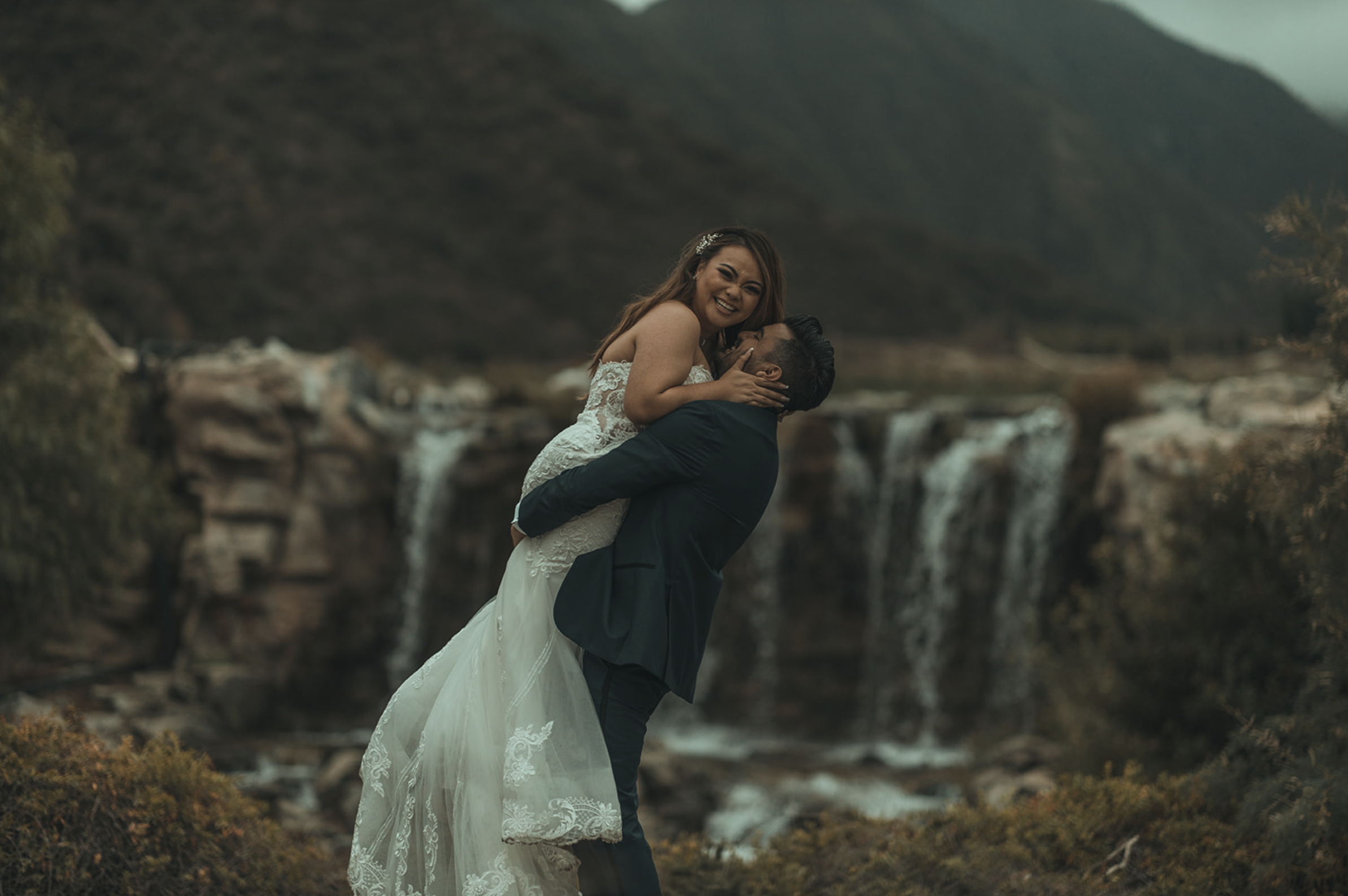 A couple dressed in wedding attire, with the man lifting the woman in his arms, smiling and embracing in front of a scenic waterfall and mountain backdrop. Glen Ivy Wedding Venue.
