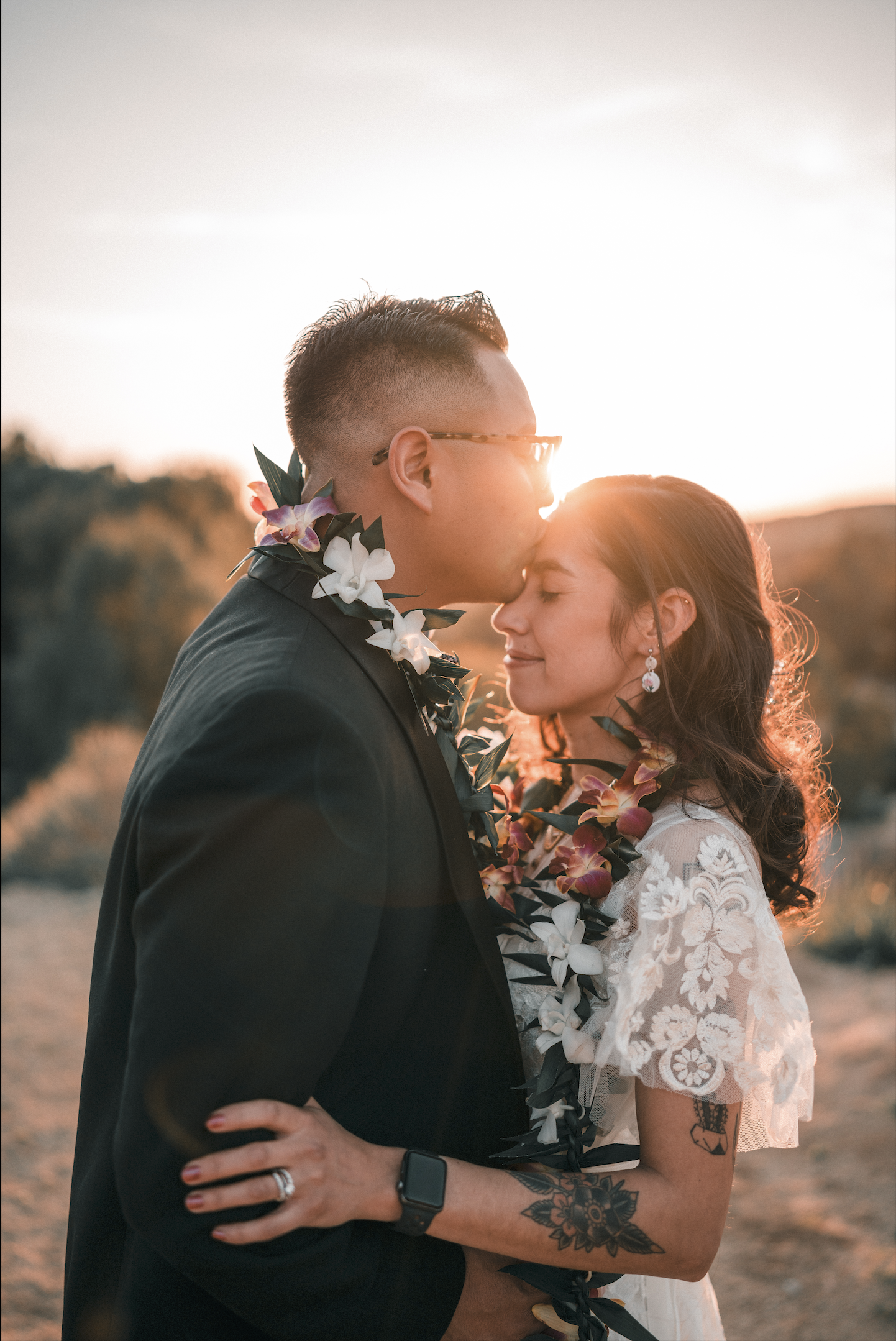 A couple on their wedding day in a sunset embrace, with the groom kissing the bride's forehead, both wearing floral leis, outdoors with a scenic landscape.