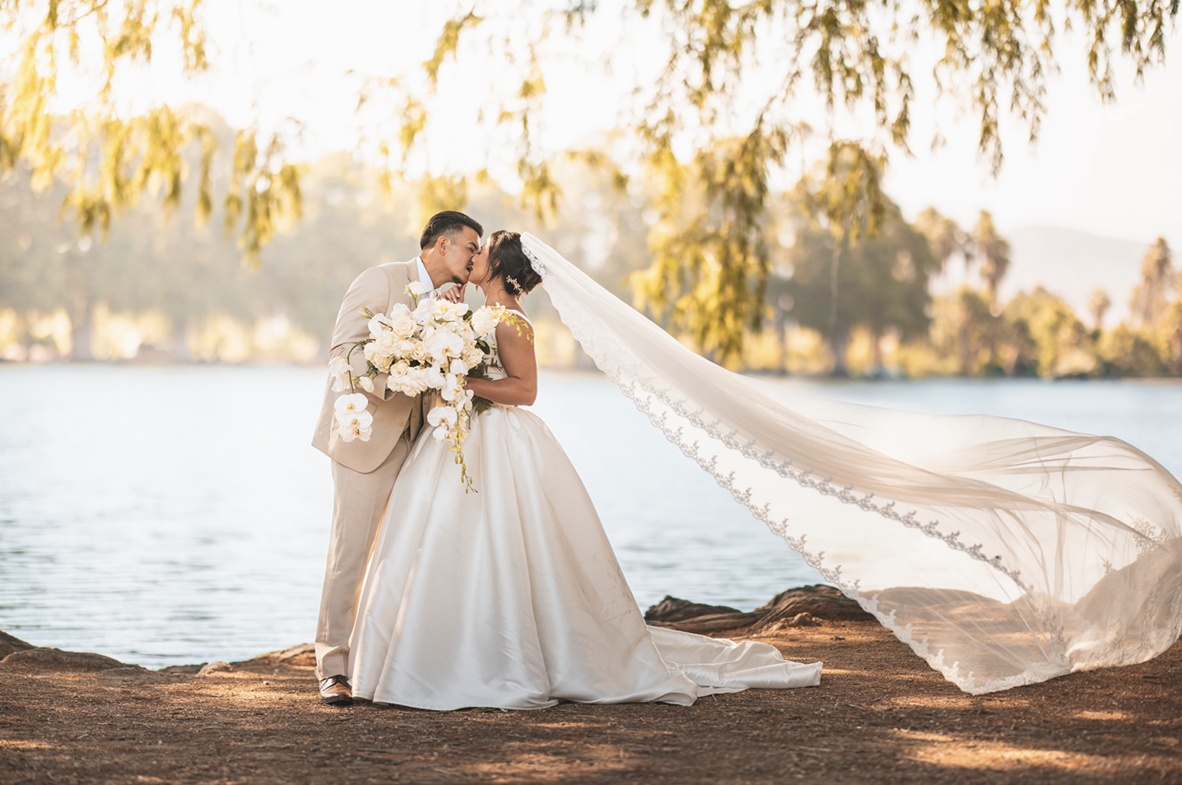 A bride and groom kissing by a lake, surrounded by trees during sunset. The bride wears a wedding gown with a long lace veil, and the groom is in a tan suit holding a large bouquet of white flowers.