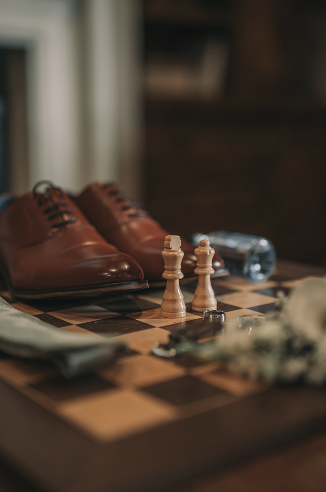 A close-up of a chessboard with two wooden pawns in focus, a pair of brown leather shoes, a water bottle, a ring, and a flower arrangement in the background. Wedgewood wedding venue Chino Hills.