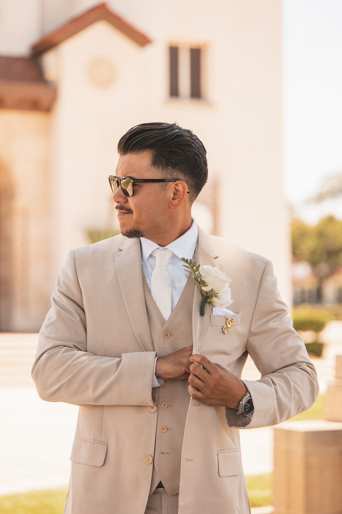 A man dressed in a beige suit with a white shirt and tie, wearing sunglasses, holding his left lapel with one hand. He has a white rose boutonniere pinned to his suit and a pin on his lapel. He is standing outdoors in front of a building with a turre
